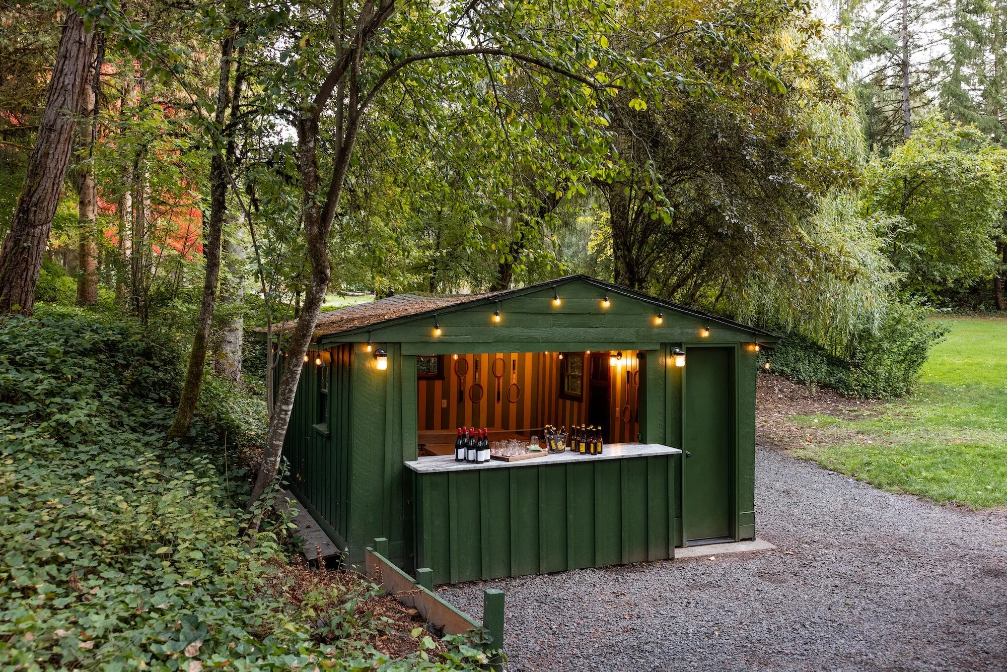 A small green shed with string lights on its roof, set within a lush wooded area. The shed's window is open, revealing a cozy interior with wine bottles and snacks on the counter. Surrounding greenery is dense with trees and bushes.