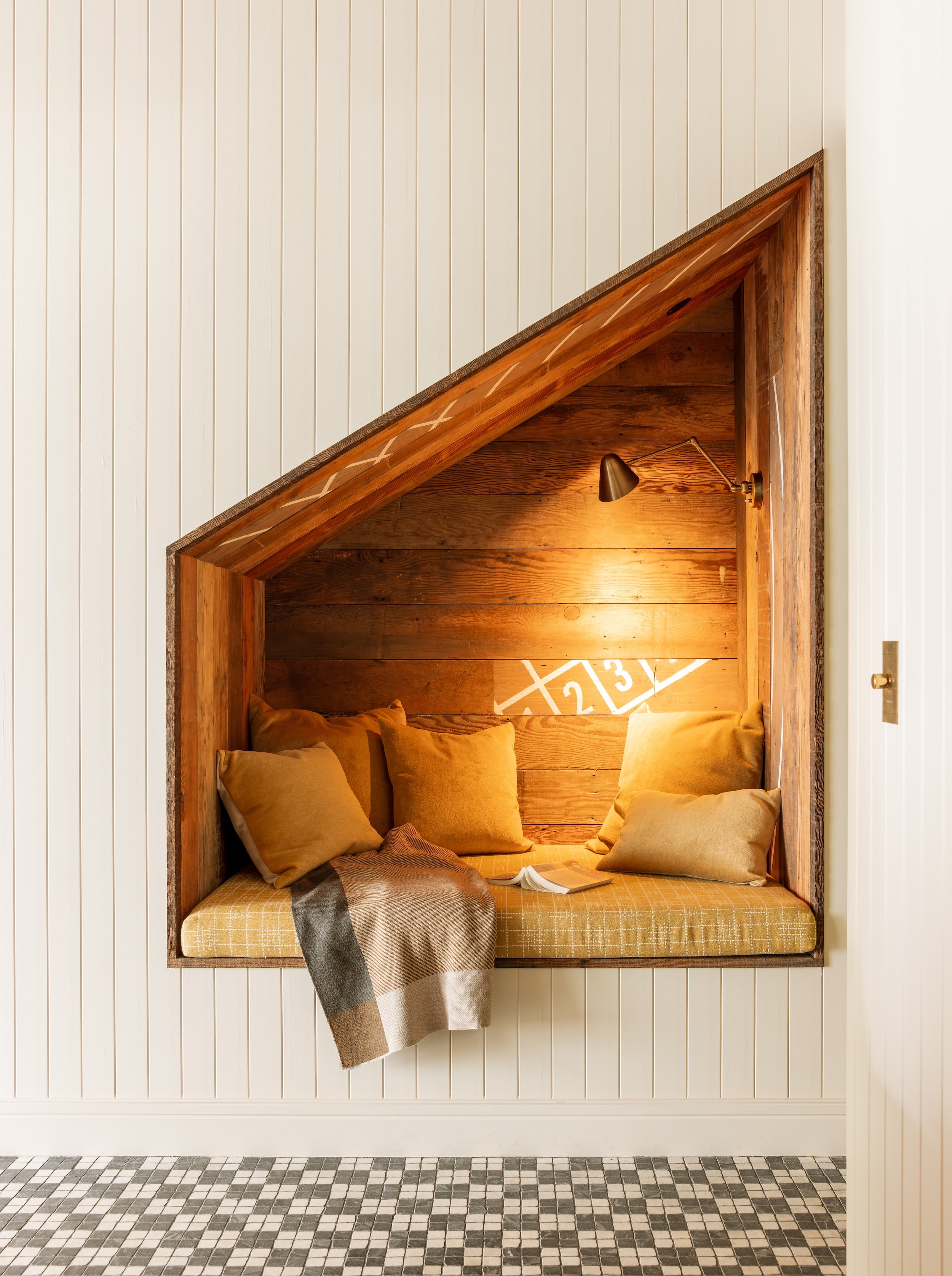 A cozy reading nook built into a wall with white vertical paneling. The nook has a wooden interior with a small reading light and is decorated with yellow cushions and a plaid blanket, with a book placed on the cushioned seat.