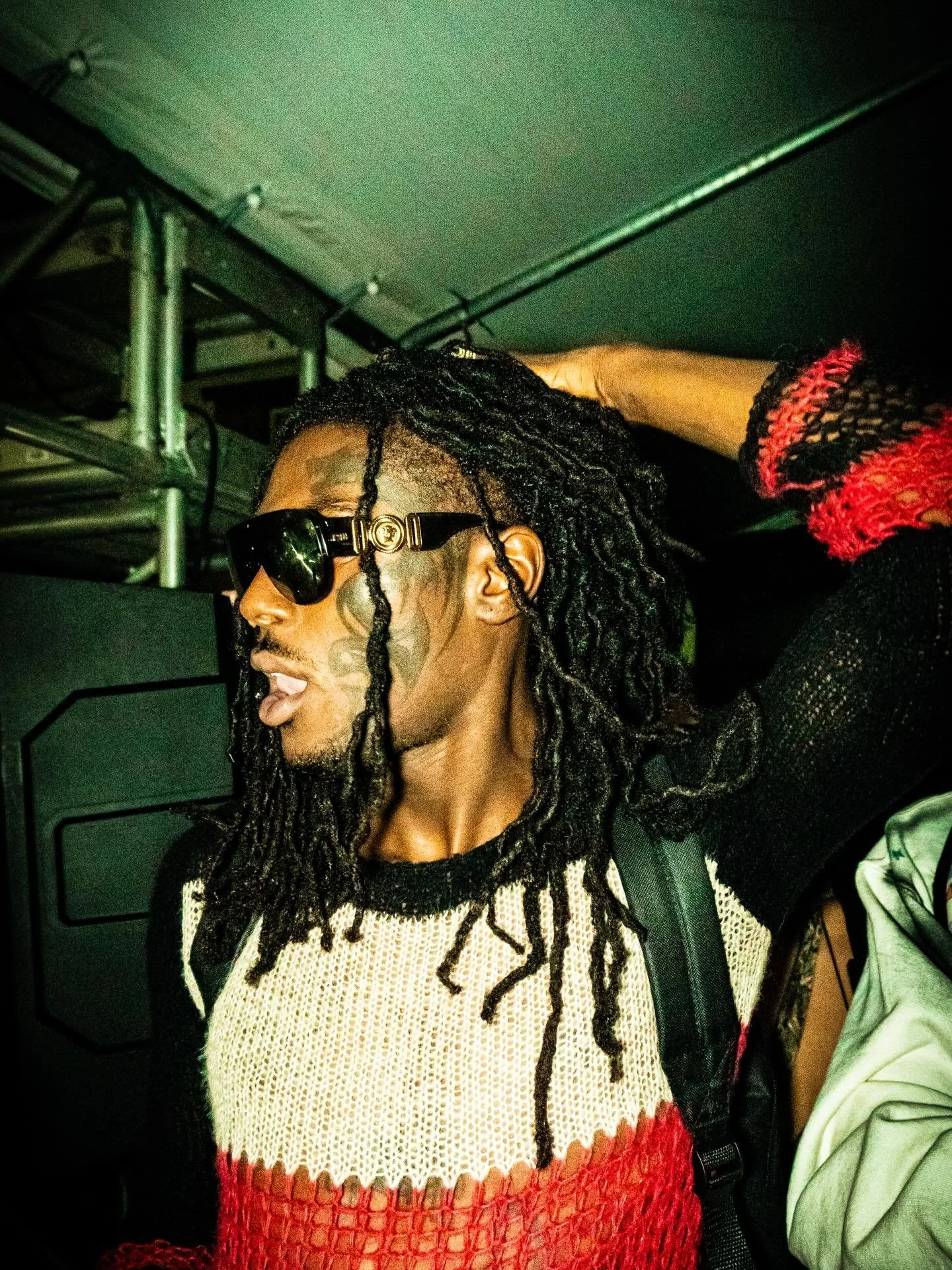 A man with long dreadlocks, wearing sunglasses with gold accents, a black shirt and a beige mesh sweater over a red garment, is adjusting his dreadlocks with his left hand. The setting appears to be indoors.