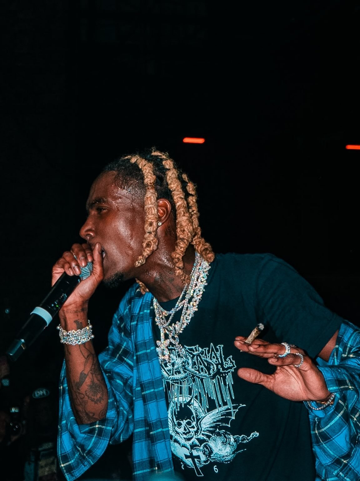 A man with blond dreadlocks holding a microphone, wearing a black T-shirt and layered jewelry, with a dark background.