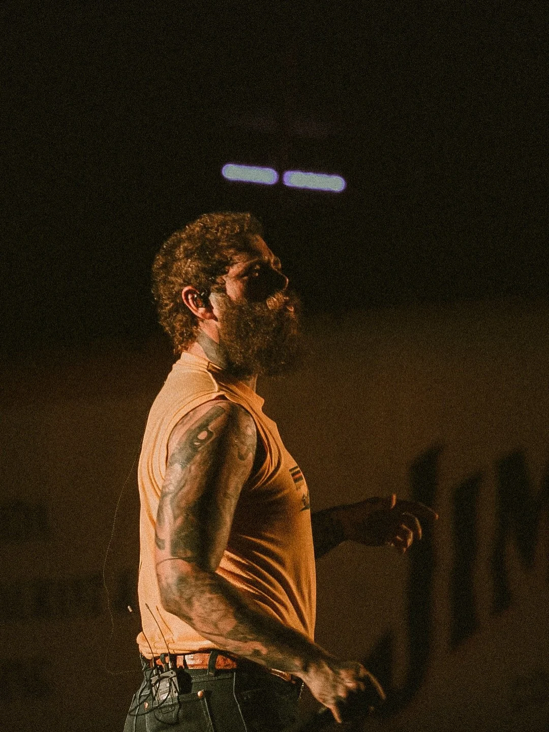 A man with a beard and curly hair, wearing a sleeveless orange shirt and black jeans, is walking in a dimly lit indoor space with dark background.