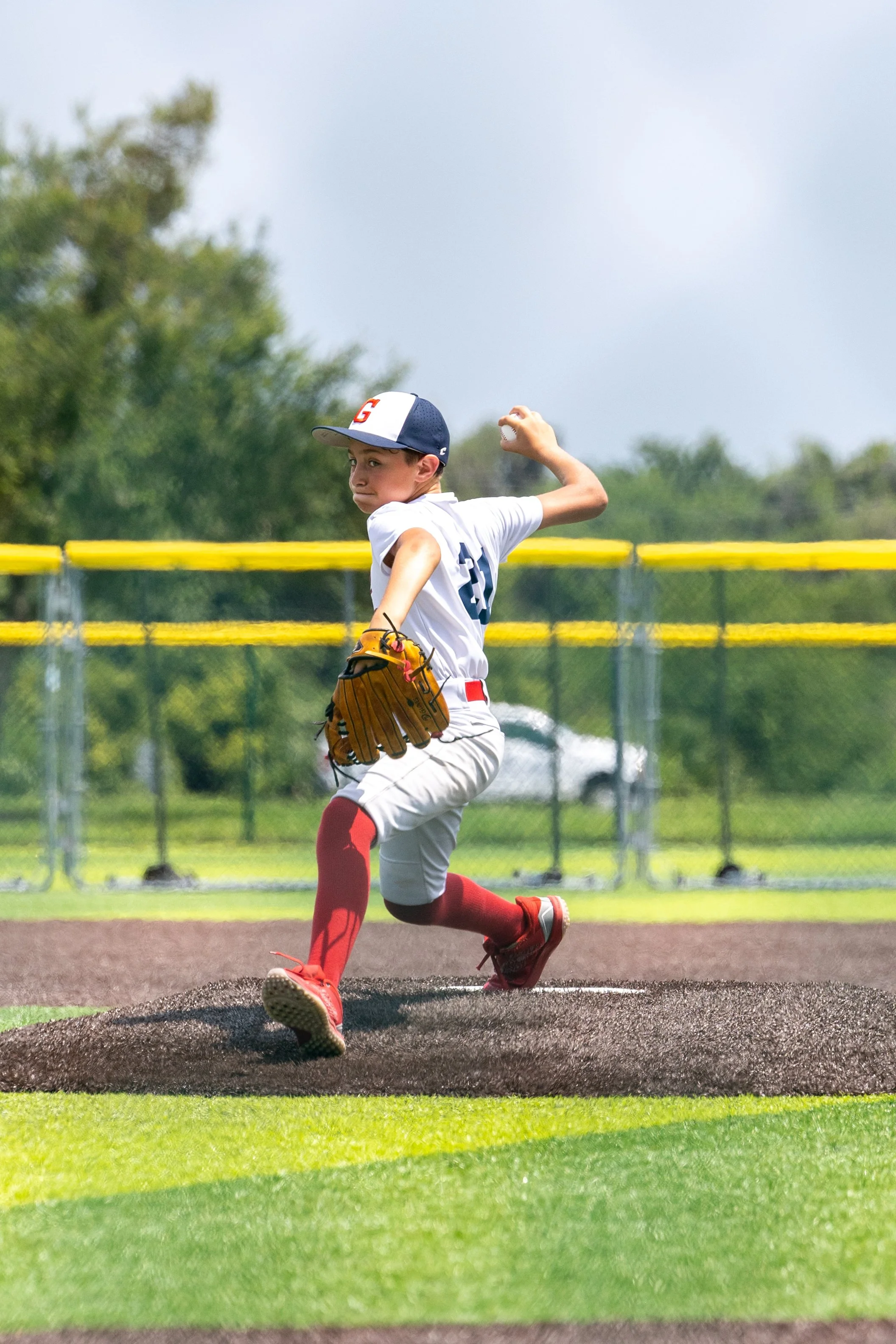 A young baseball player in uniform and red socks is sliding into a base on a baseball field during daytime, with a yellow fence and trees in the background.