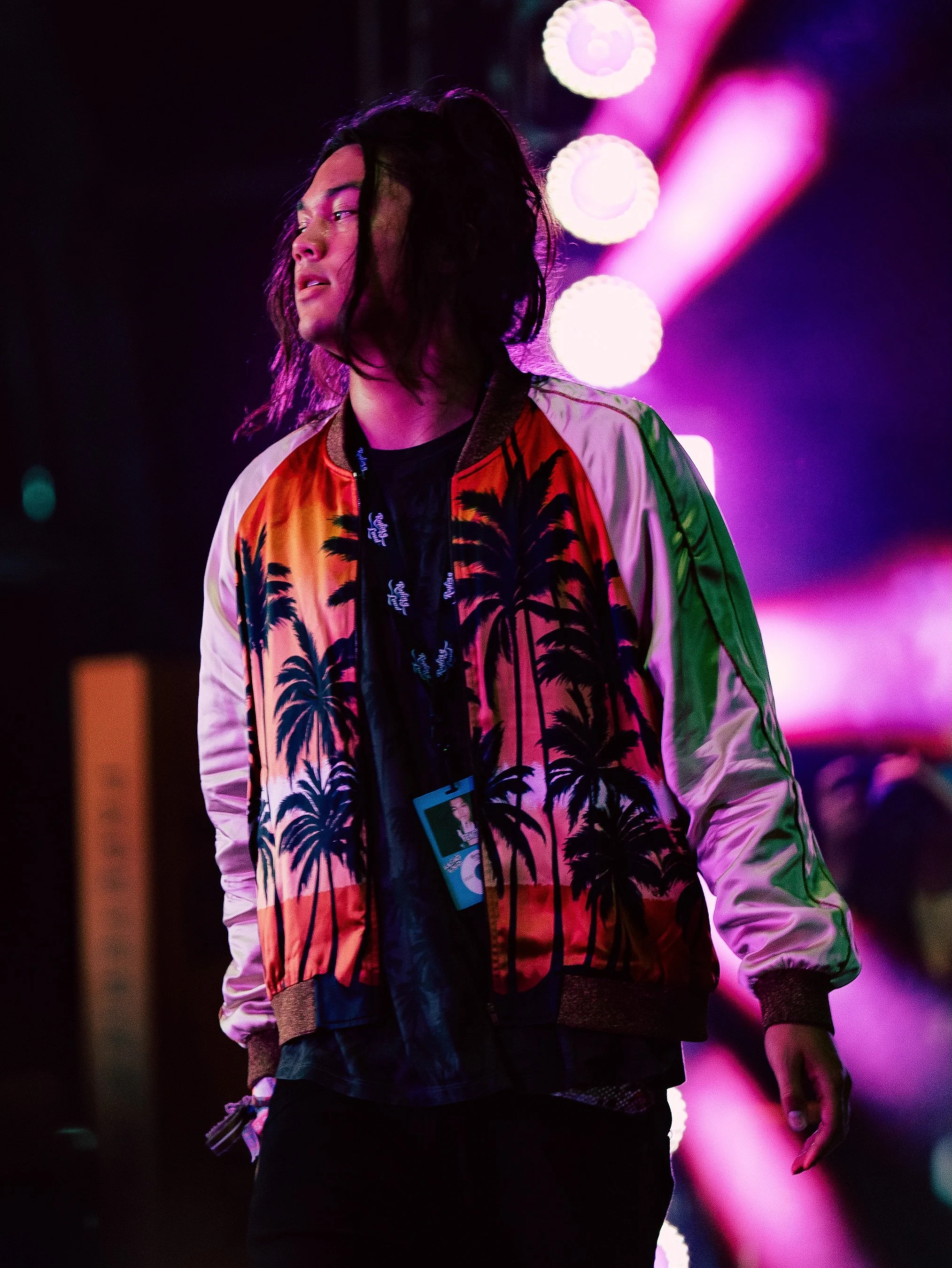 A person with shoulder-length dark hair stands against a background of pink and purple stage lights, wearing a colorful jacket with palm tree patterns and a black shirt underneath.