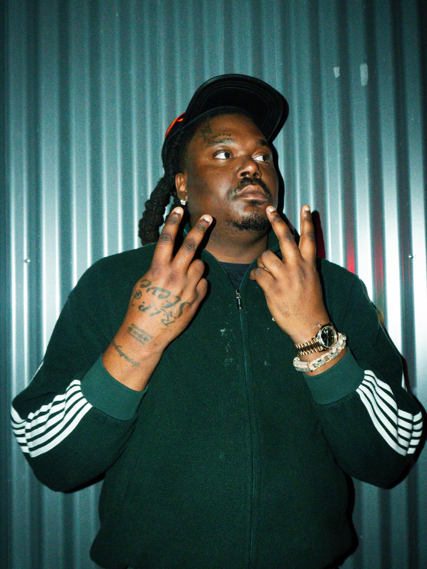 A man with braided hair and tattoos on his hands is wearing a black jacket with white stripes on the sleeves, a black cap tilted backward, and gold jewelry, standing against a metallic corrugated background.