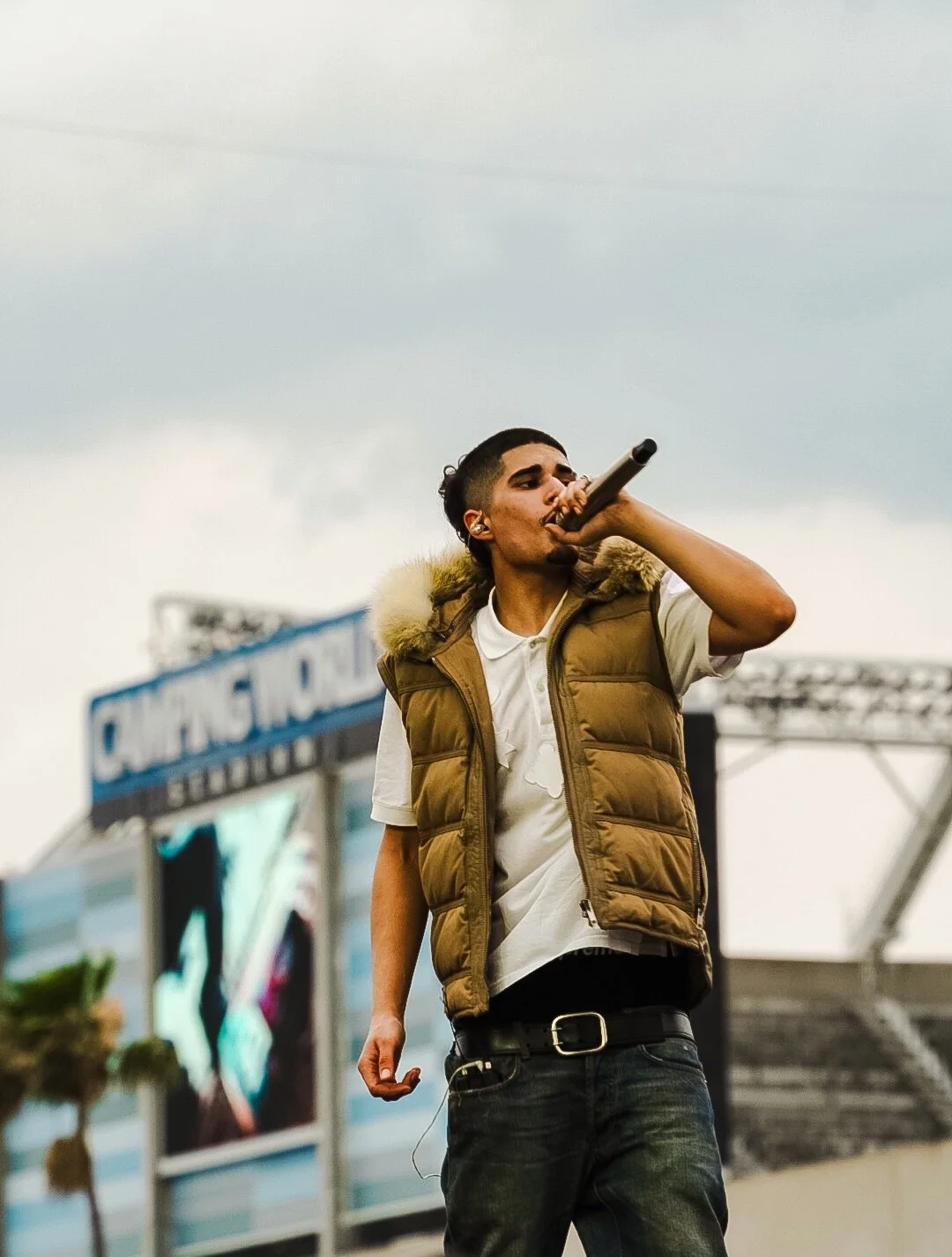 A young man performing at an outdoor event, holding a microphone, with a sign in the background that says "Champs Sports".