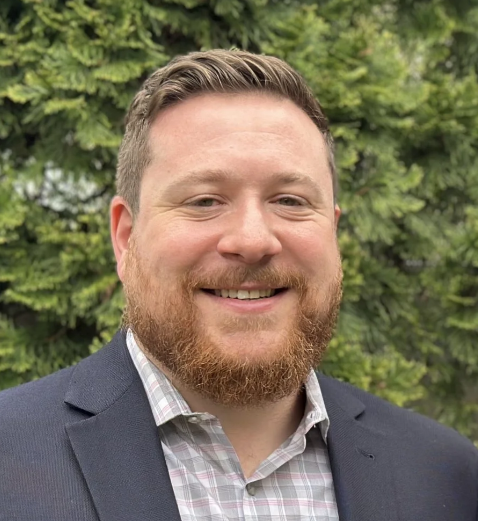 A smiling man with a beard wearing a dark suit jacket and a checkered shirt, standing outdoors in front of green trees.