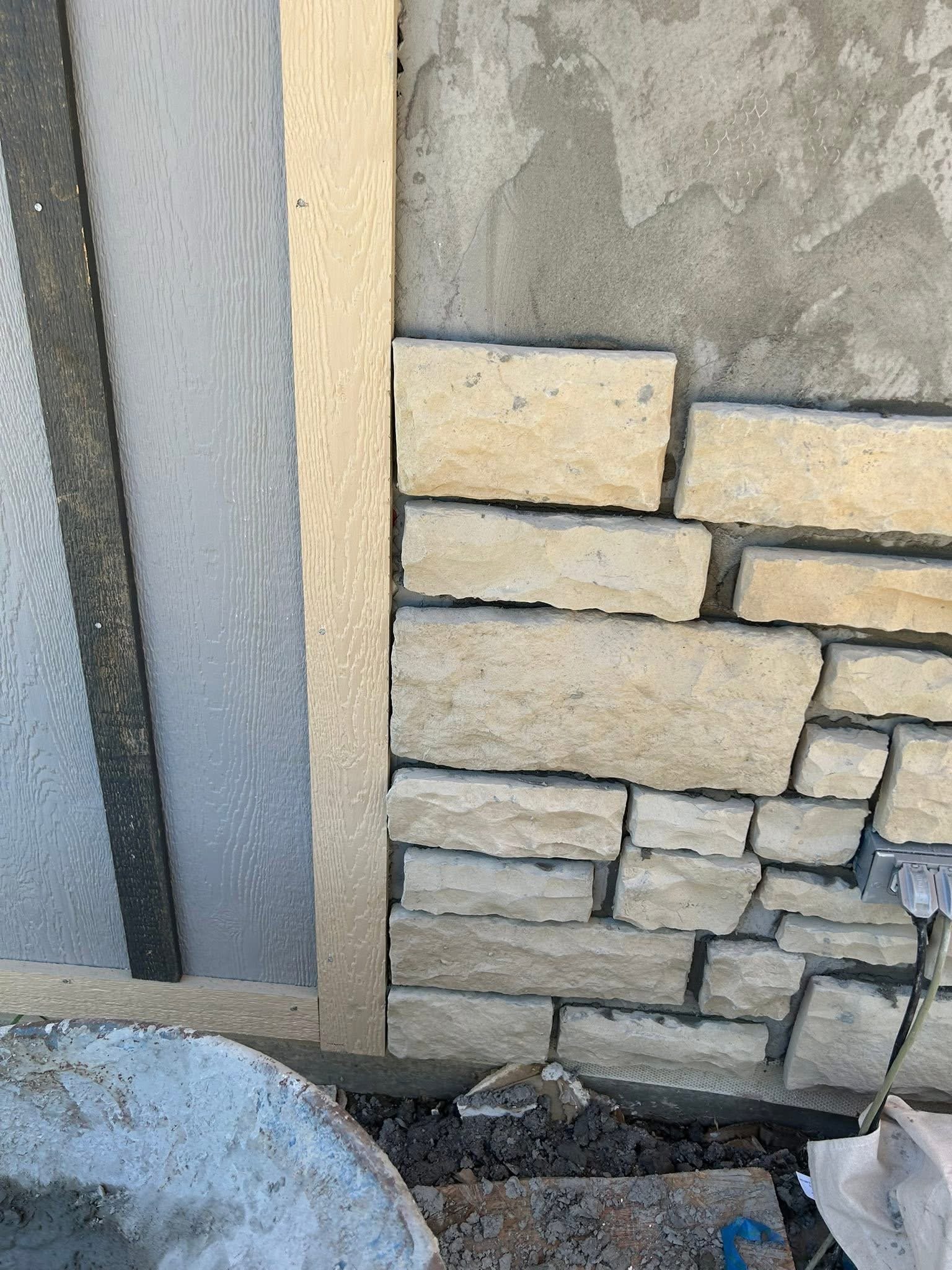 Close-up view showing a section of a wall with beige stone bricks, gray mortar, and adjacent wooden siding painted in gray and natural wood color.