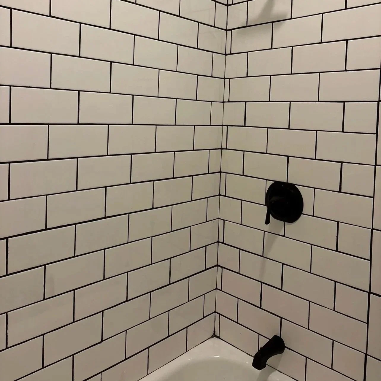 Corner of a shower with white subway tiles, black grout, a black shower valve, and a black diverter.