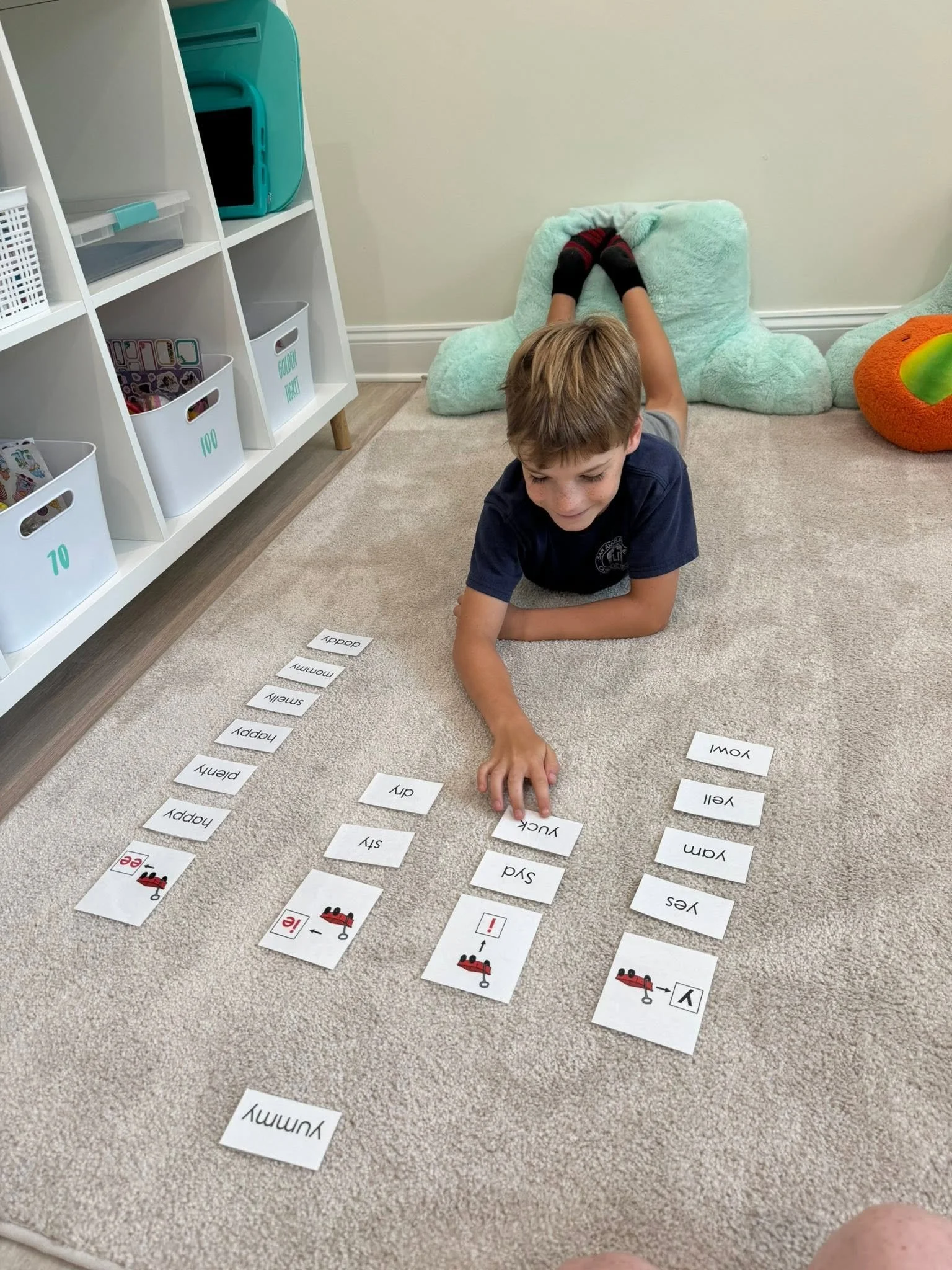 A young boy is lying on the carpeted floor, reaching out to arrange alphabet cards with words and images related to emergency services, such as 'ambulance,' 'fire,' and 'help.' There are storage shelves with bins and a plush armchair in the background.