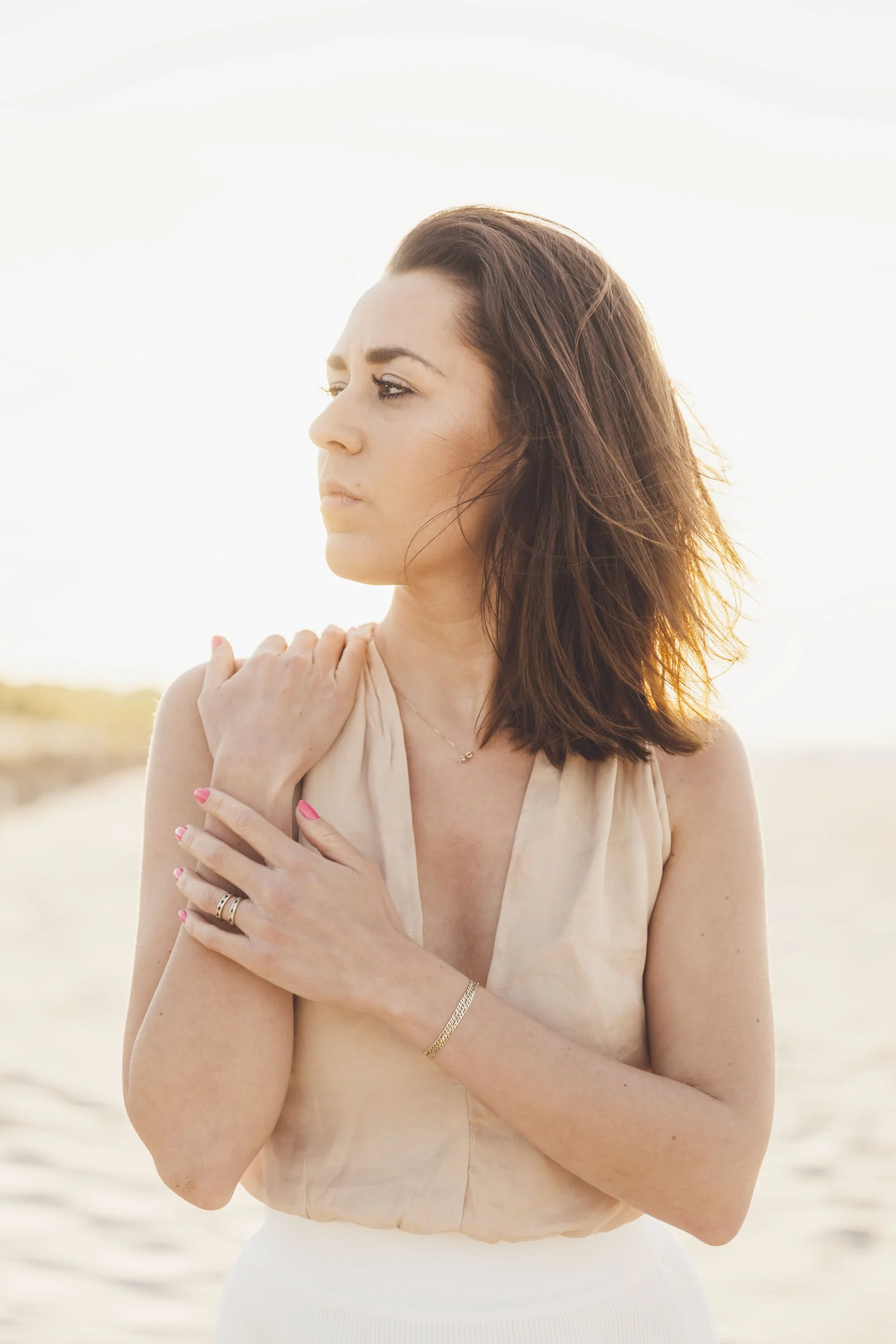A woman with brown hair and light skin poses outdoors, crossing her arms over her chest and gently holding her shoulders, with sunlight illuminating her face and a blurred background of a sandy area.
