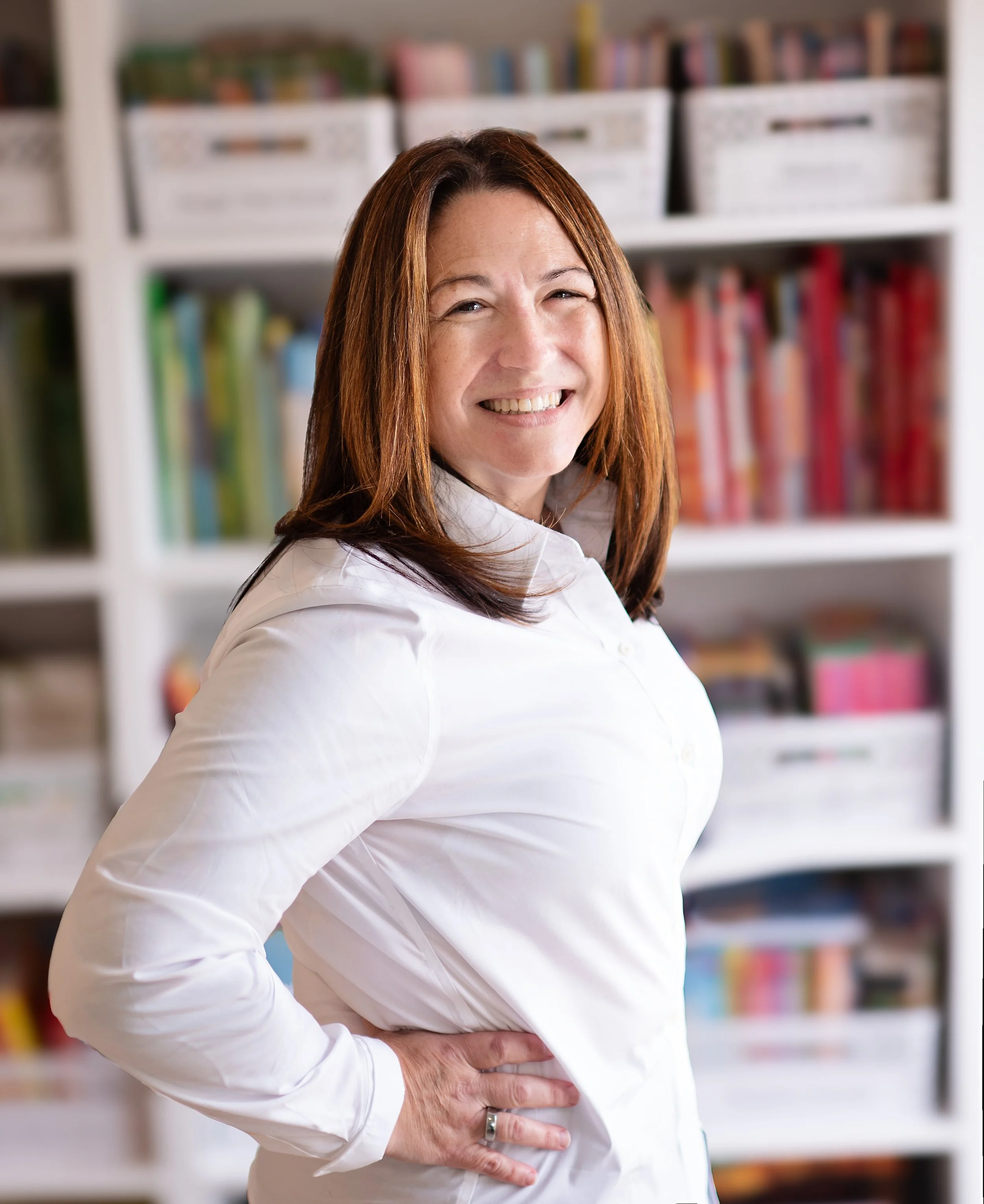 Ms. Heather wearing a white blouse, smiling with her hand on her hip, in front of a bookshelf filled with books.