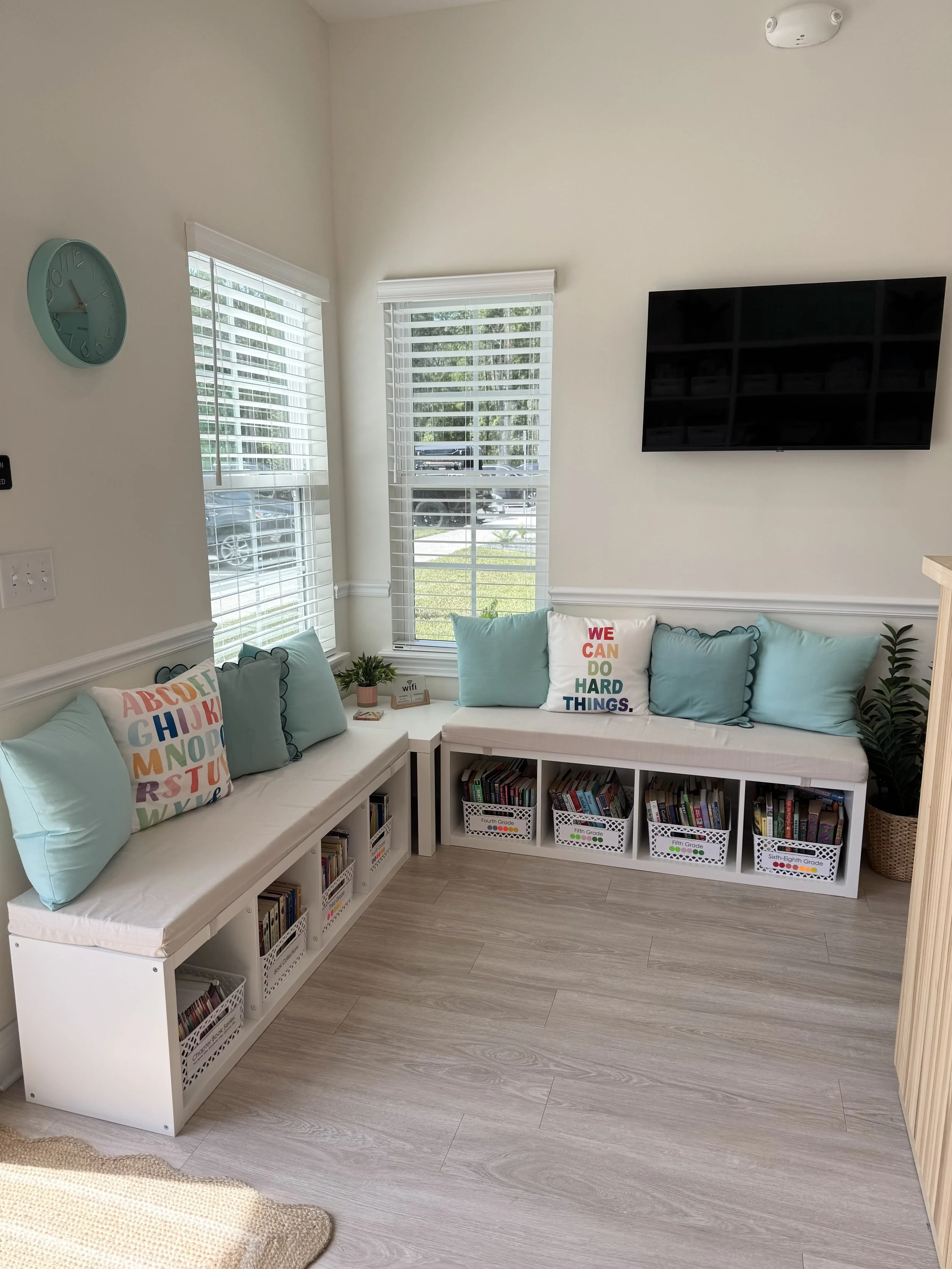 A cozy seating area with light teal cushions and a decorative pillow that says "We Can Do Hard Things," next to a window with white blinds, a wall-mounted TV, and a bookshelf filled with children's books, with a small potted plant on the side table.