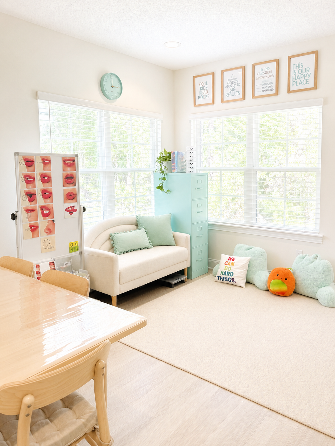 Bright classroom corner with large windows, colorful wall art, a small cream sofa with pastel pillows, a light blue filing cabinet, a plush animal pillow, and a whiteboard with face lips illustrations.