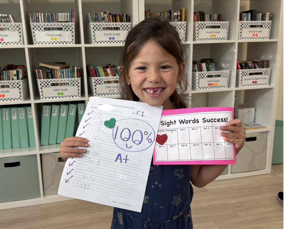 A young girl smiling and holding up her perfect score sheet and handwritten work in a classroom or library with shelves of books in the background.