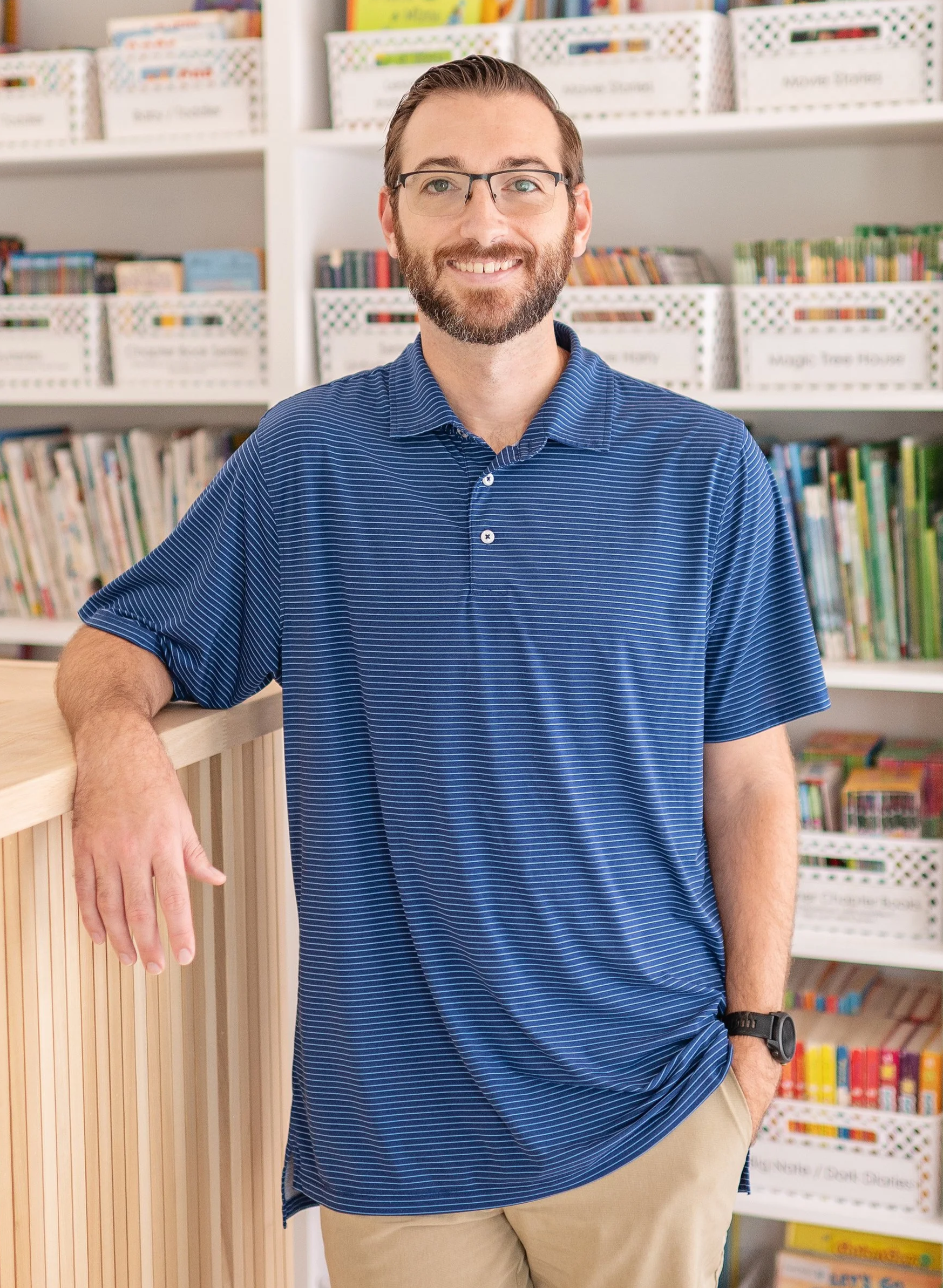 Mr. Mike wearing a blue striped polo shirt and khaki pants, standing in front of shelves filled with books and magazine boxes in a library or bookstore setting.