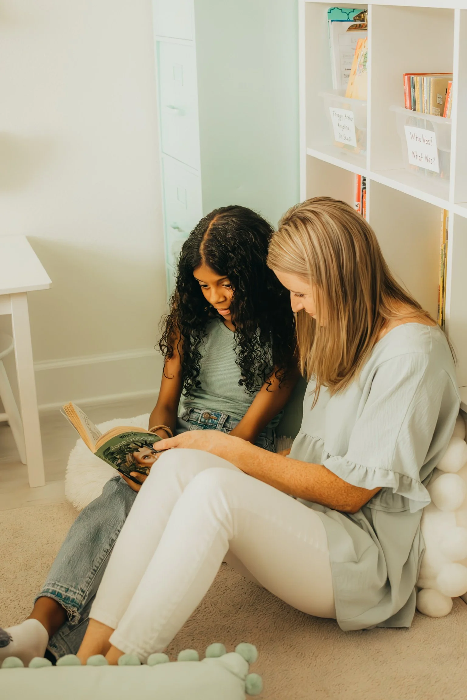 Emily Lavery and a student sit on the floor and look at a magazine together in a cozy, well-lit room with cream-colored walls and a white bookshelf. The bookshelf contains books and labeled bins.