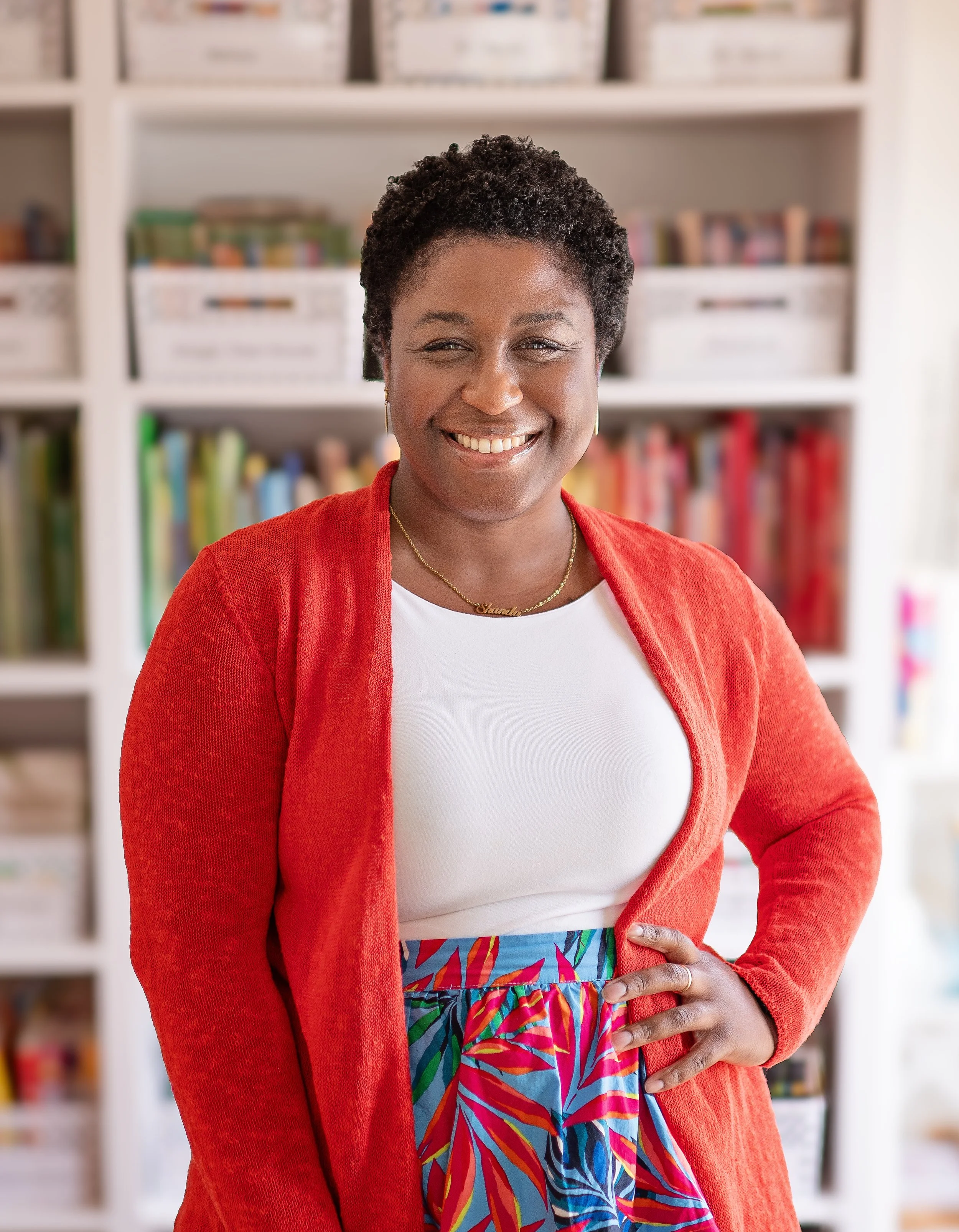 Ms. Shanda wearing a red cardigan, white top, and a colorful skirt, standing in front of a bookshelf filled with books.