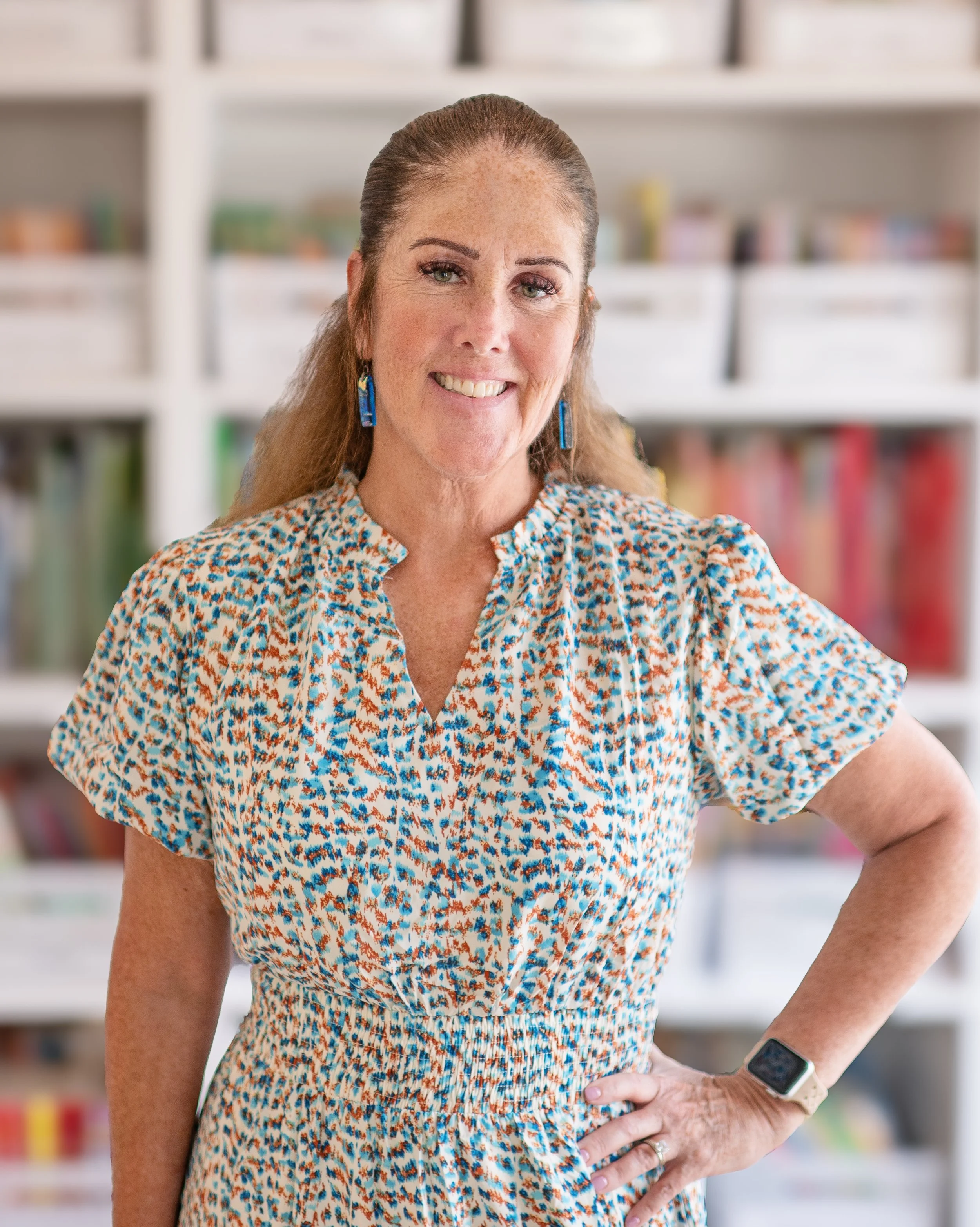 Mrs. Jen, wearing a colorful floral dress with a V-neckline and puffed sleeves, stands with her right hand on her hip in front of a bookshelf with various books. She is smiling gently and wearing blue earrings and a smartwatch on her left wrist.