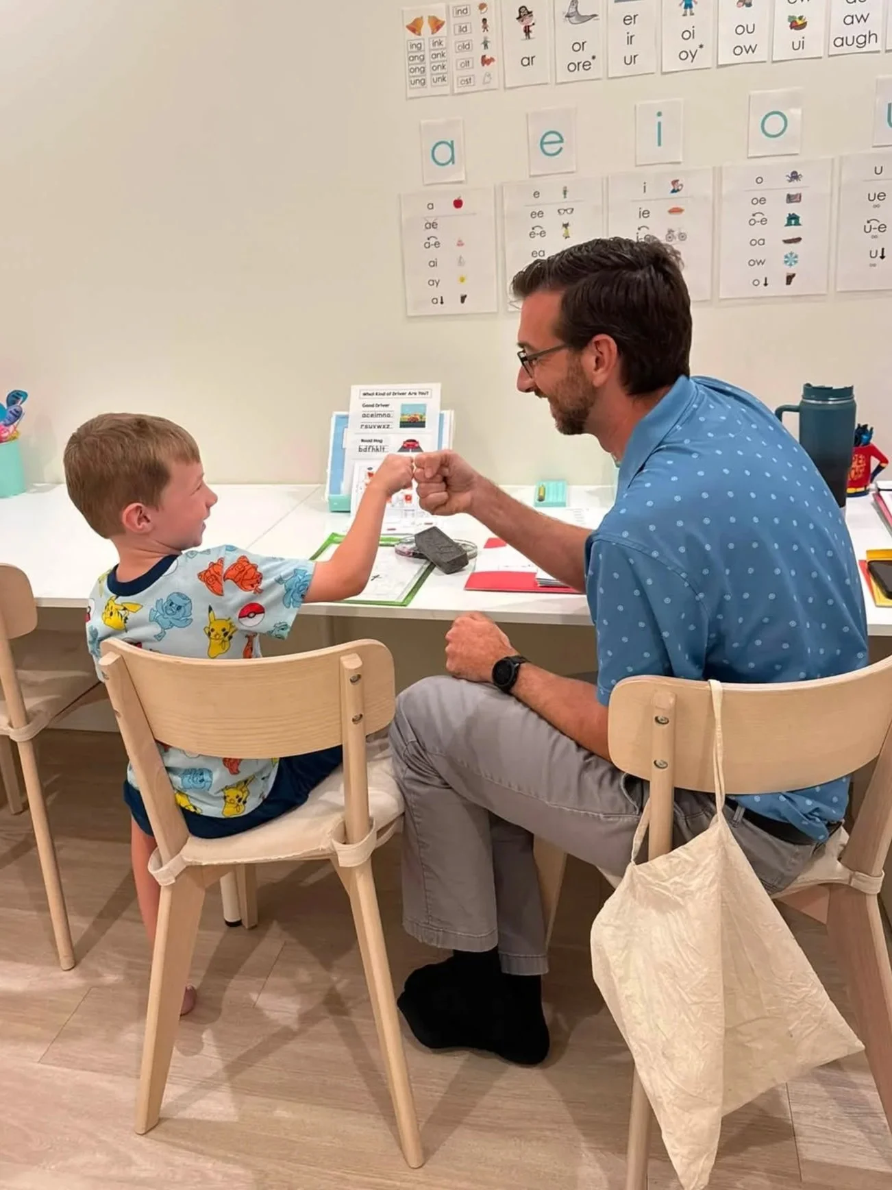 A student and teacher are sitting at a table in a classroom, engaging in a fist bump. The boy is wearing a colorful Pokémon t-shirt, and the man is wearing a blue polka-dotted shirt and glasses. They are smiling at each other.