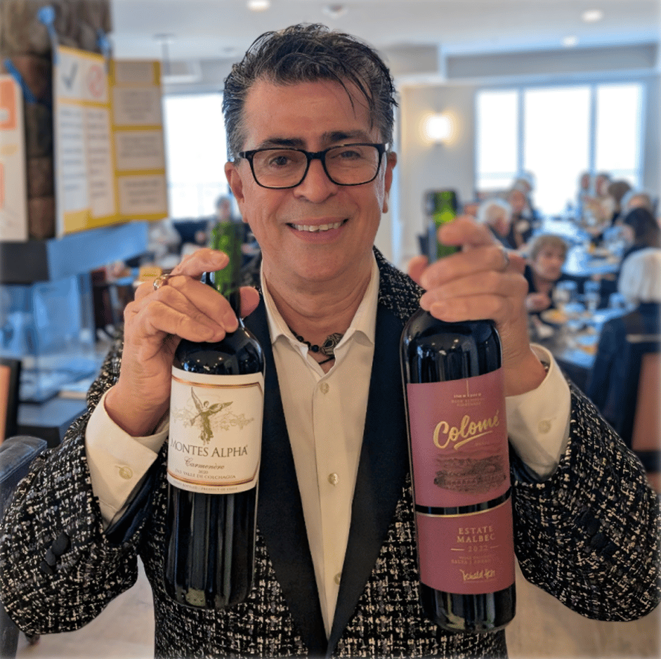 Rico smiling as he holds two bottles of red wine (a Carmenere and a Malbec) at a wine tasting.