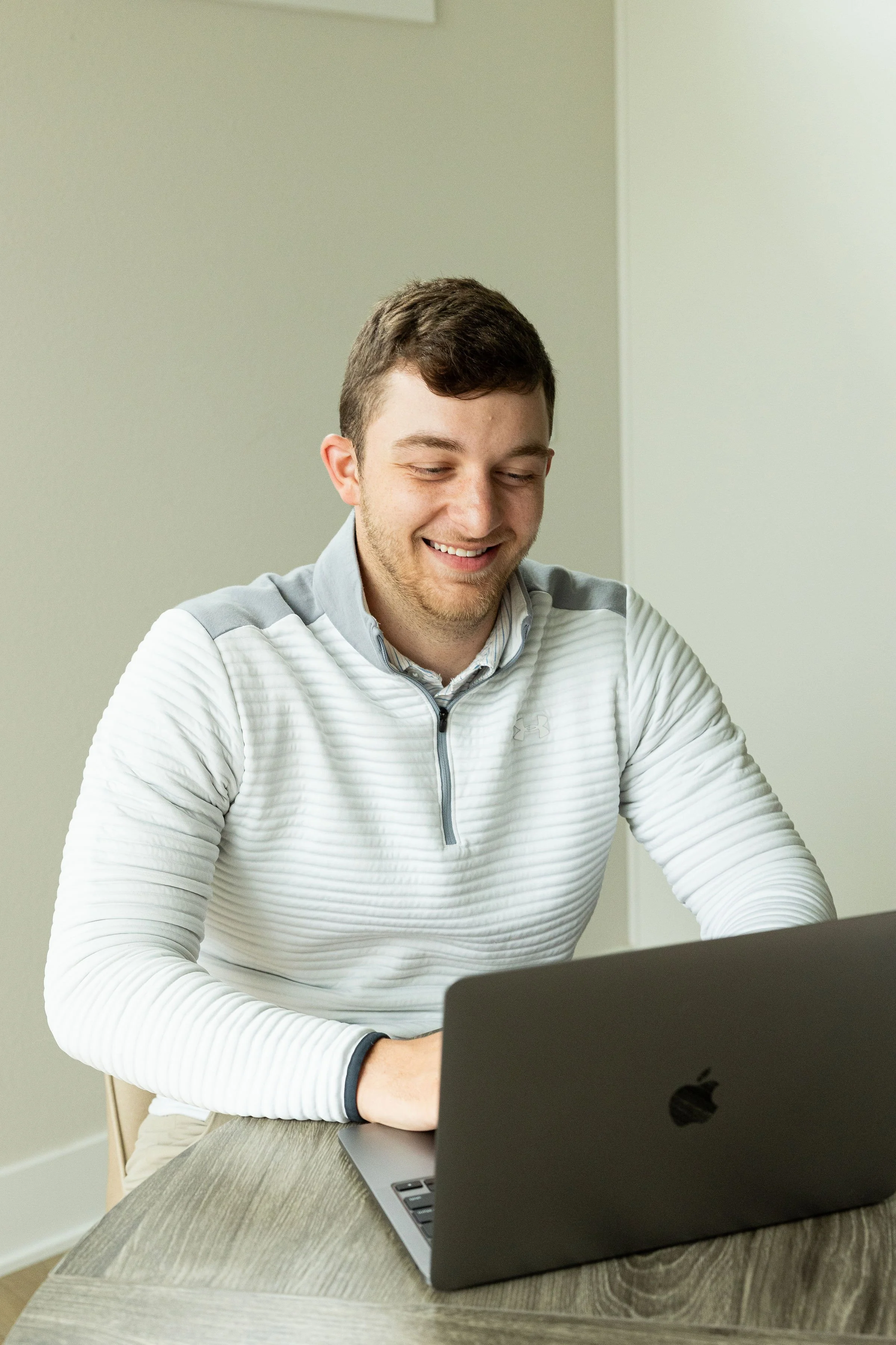 A young man with short brown hair sitting at a wooden table, smiling while using a black laptop, wearing a white zip-up jacket with gray accents.