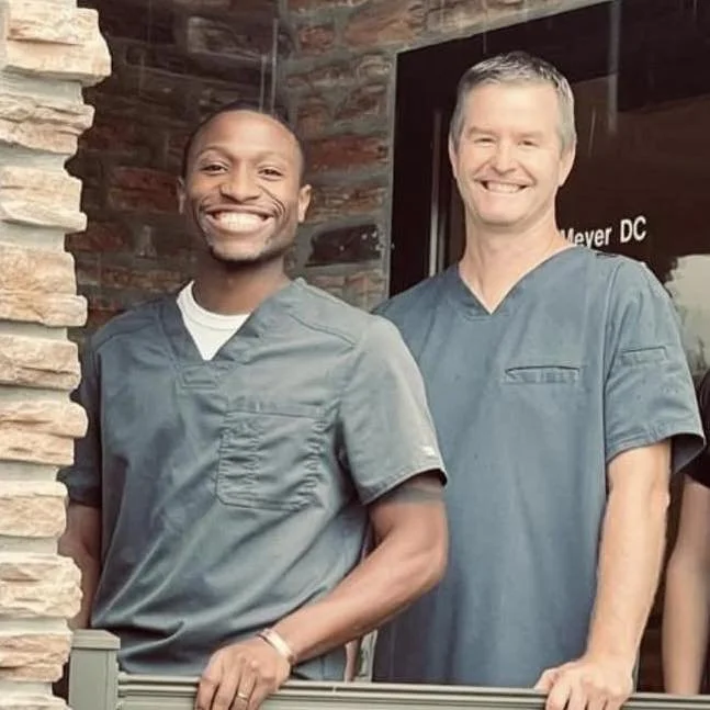 Two smiling medical professionals in scrubs standing outside a building with a stone wall and a sign that reads 'Meyer DC'.