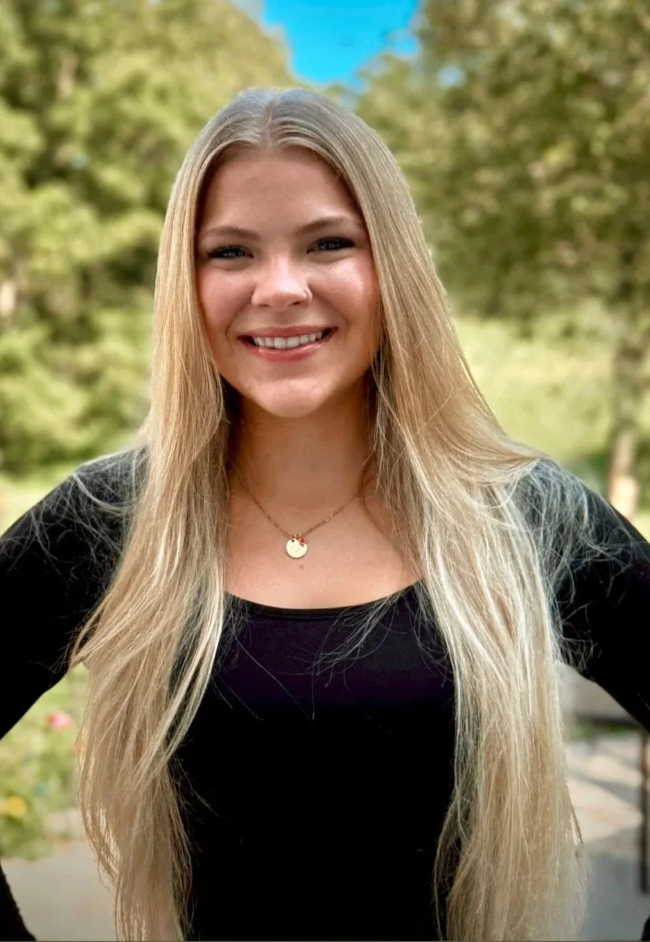 A young woman with long blonde hair smiling outdoors with green trees and a blue sky in the background.