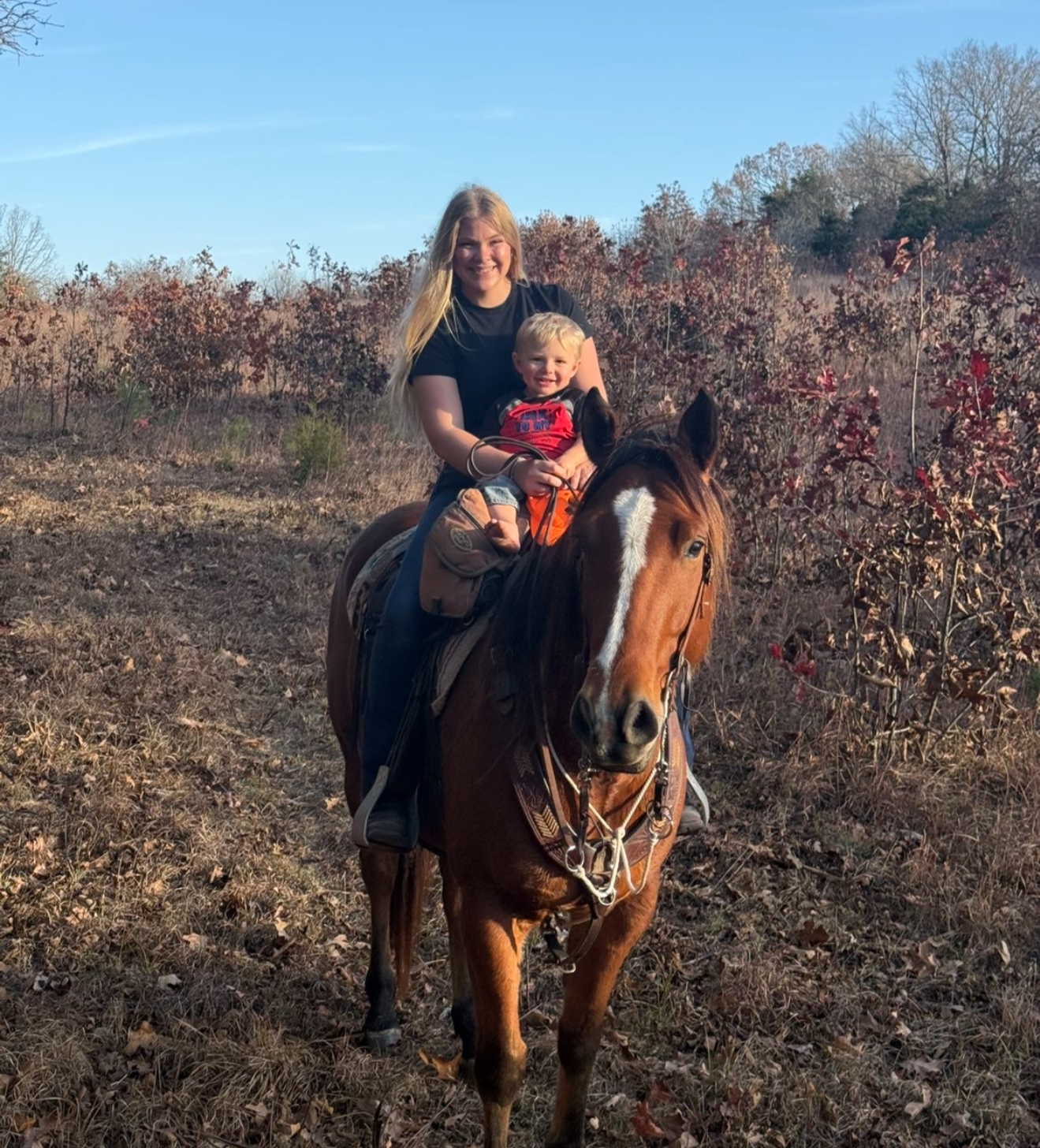 A woman and young boy riding a brown horse on a trail through a wooded area with autumn foliage, clear blue sky in the background.