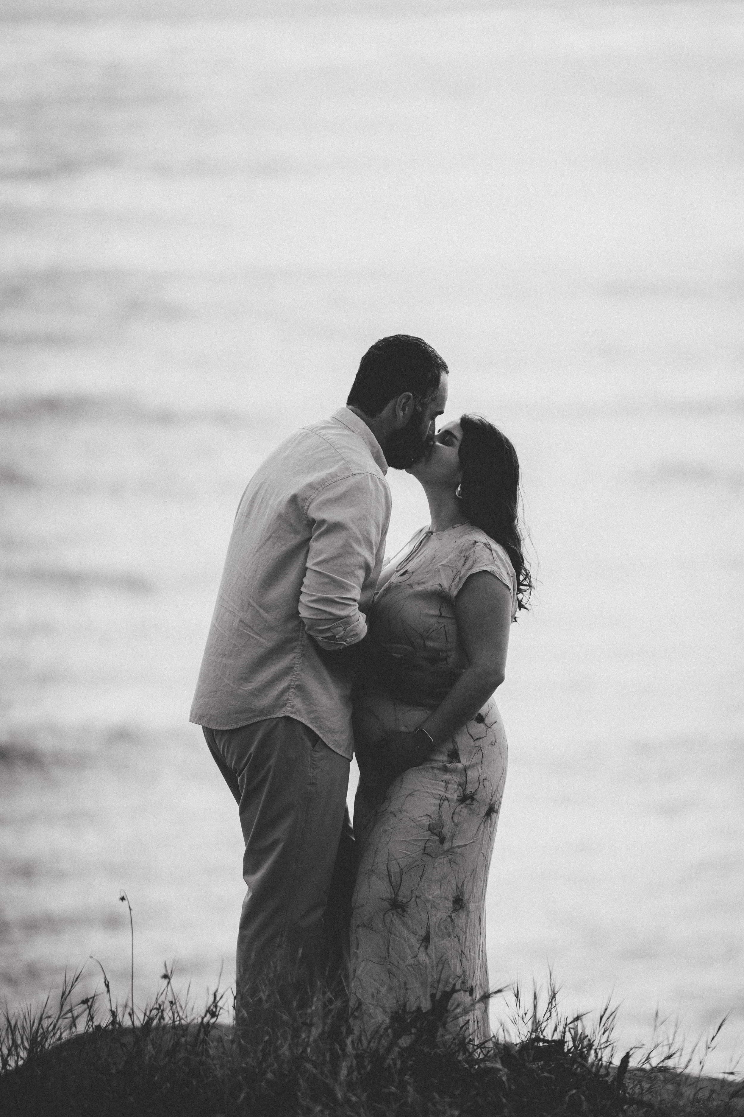 A black and white photo of a couple standing closely on a grassy area by the water, sharing a kiss.