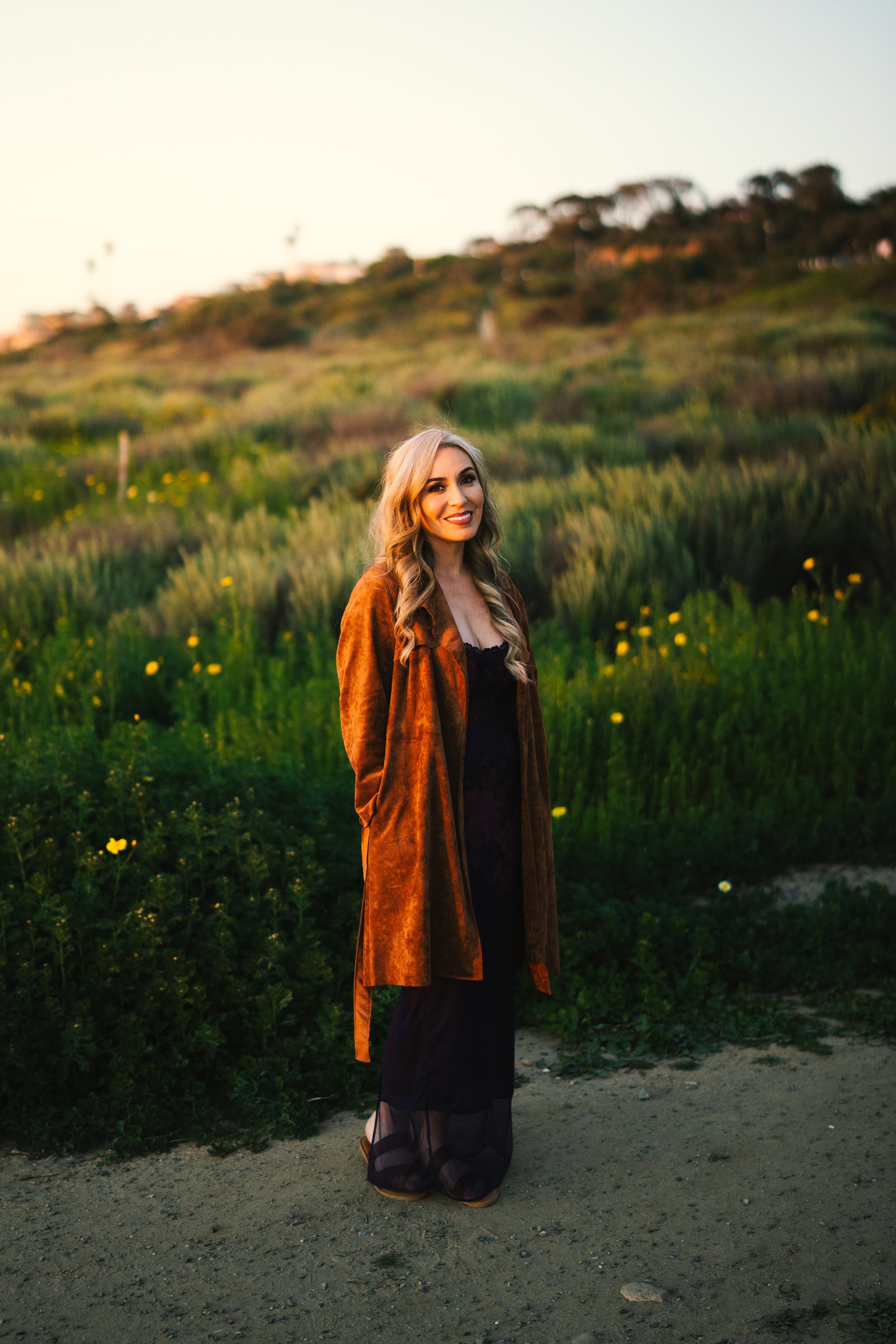 A woman standing outdoors on a dirt path with green grass and yellow flowers, smiling at the camera, with a hillside and trees in the background during sunset.
