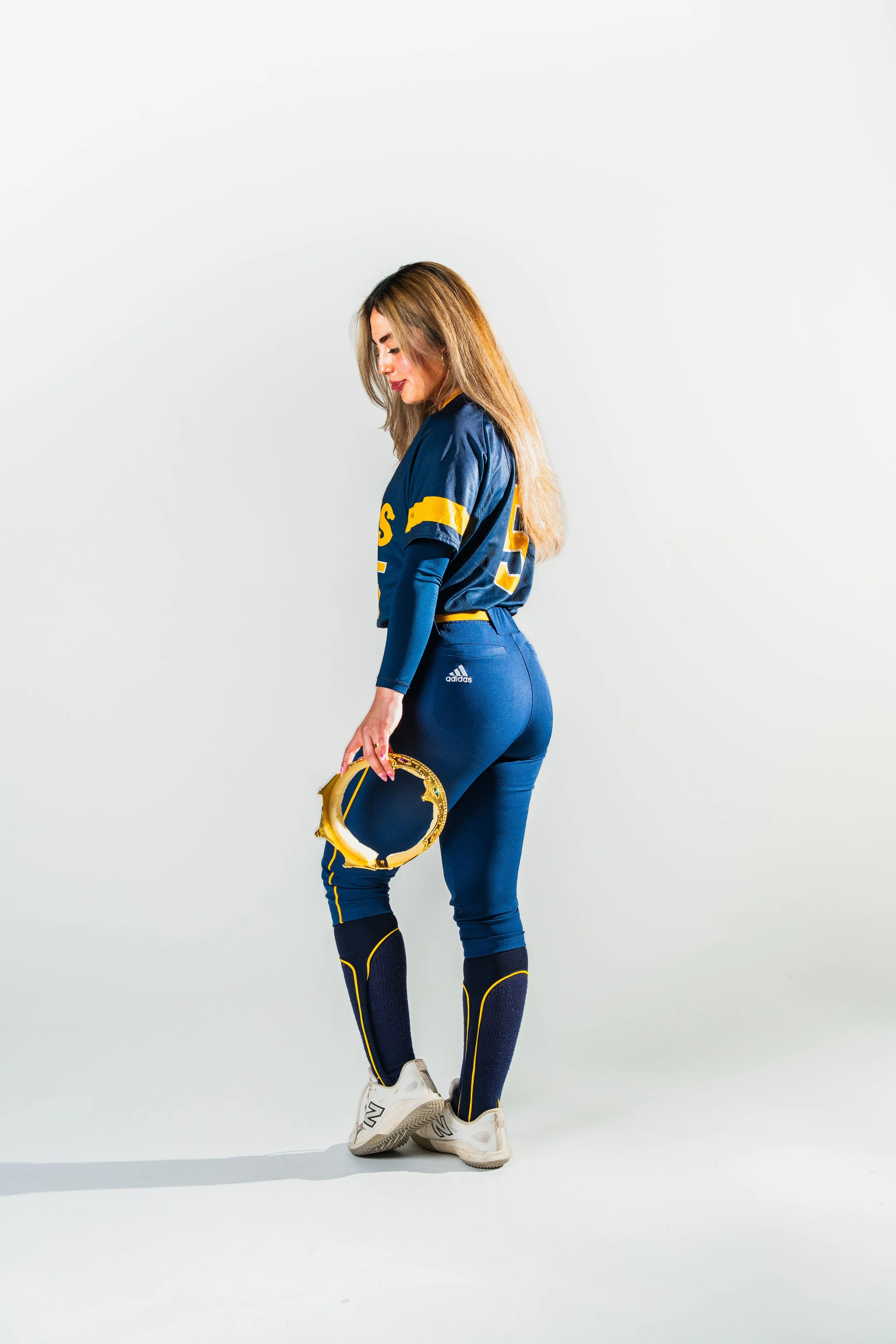 A woman in this image is wearing a blue sports uniform with yellow accents, holding a gold-colored crown, and standing against a plain white background.