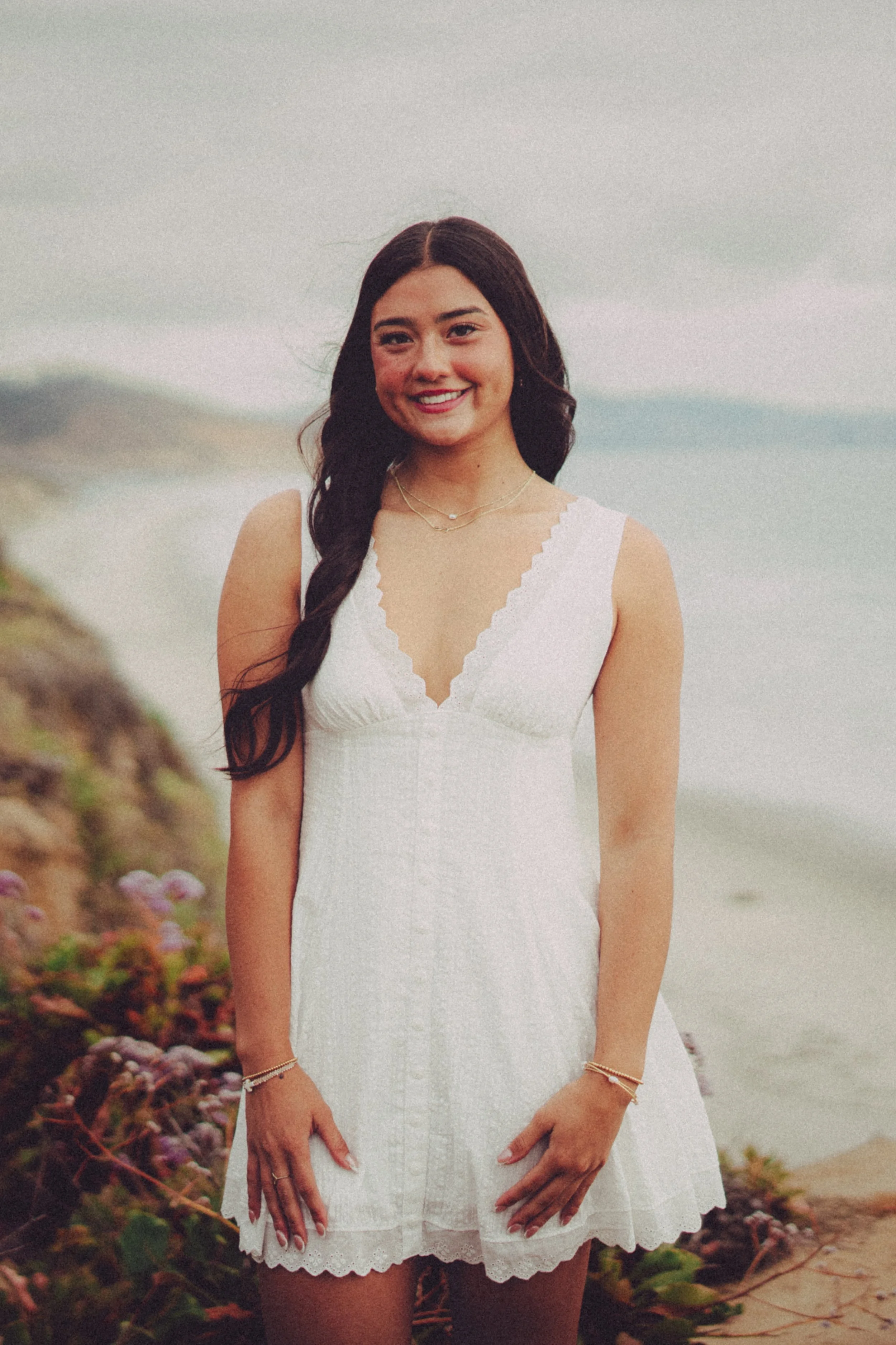 A young woman with dark wavy hair and a white dress standing on a beach with rocks and plants in the foreground and ocean in the background.