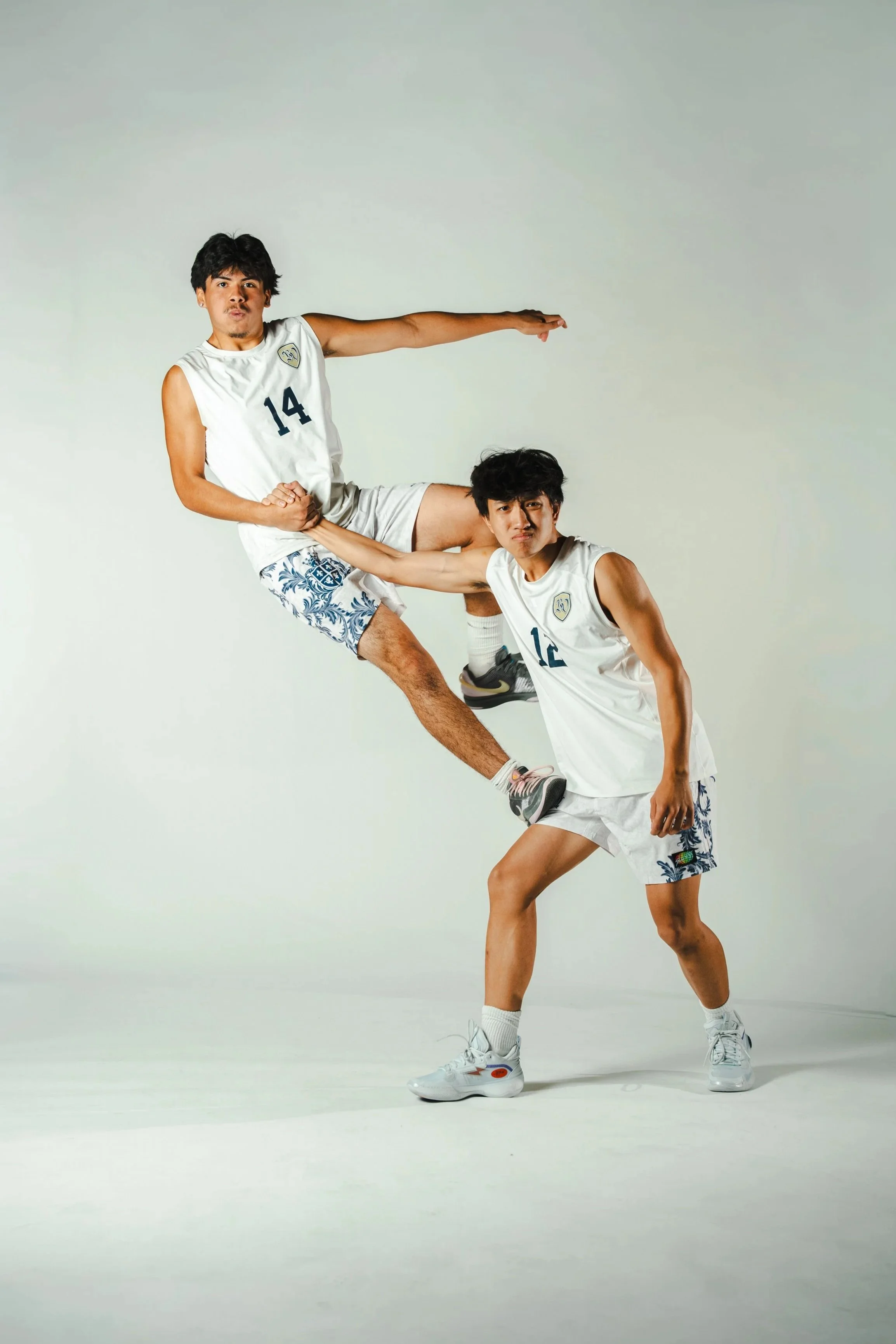 Two young men in athletic wear striking a synchronized pose against a plain white background. One is standing with one leg raised and bent, holding a hand on his knee, while the other balances on one foot with his arm extended, both looking at the camera.
