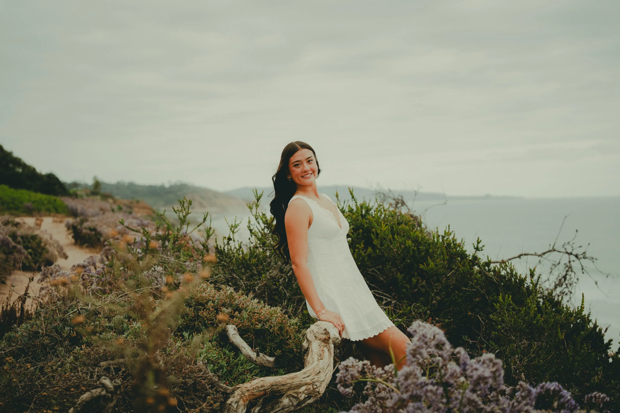 A young woman in a white dress smiling while sitting on a driftwood log among coastal vegetation, with the ocean and cloudy sky in the background.