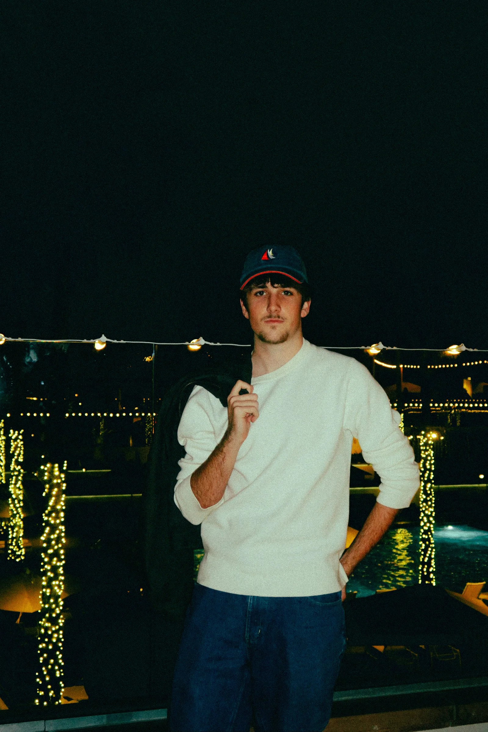A young man with light skin, dark hair, and a short beard standing outdoors at night near string lights, wearing a white long-sleeve shirt, dark jeans, and a baseball cap with a sailboat logo, holding a jacket over his shoulder.