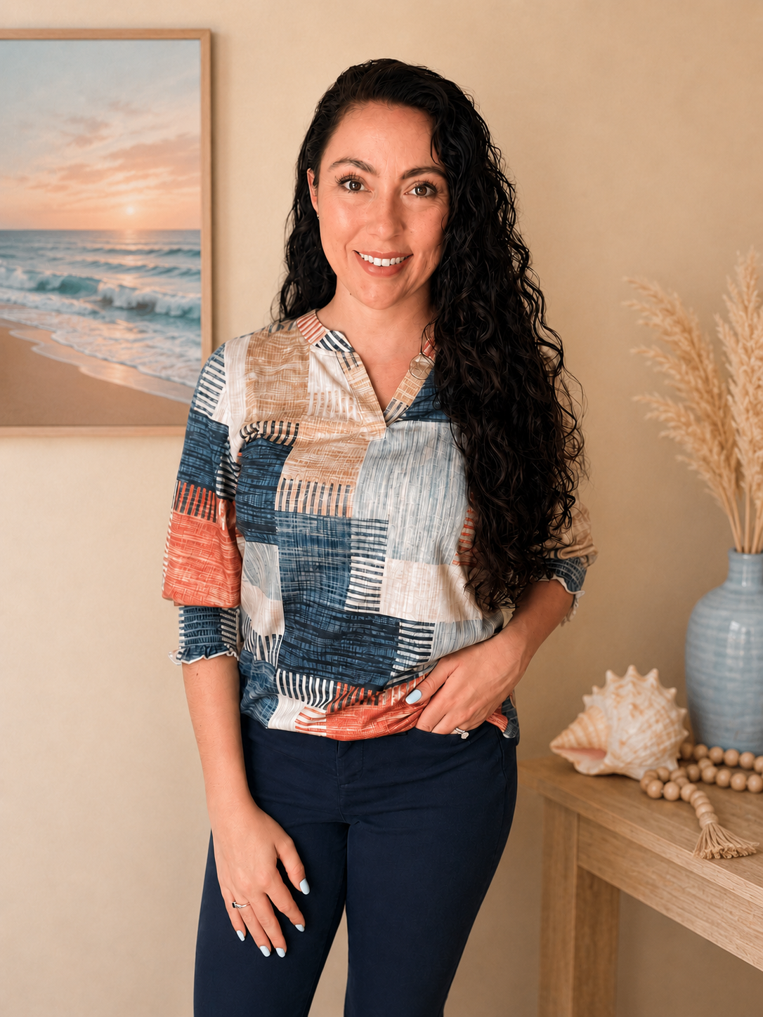 A woman with long, curly black hair, wearing a multicolored patchwork blouse and dark pants, standing in a room with beach-themed decor including a picture of a sunset over the ocean, seashells, and dried plants.