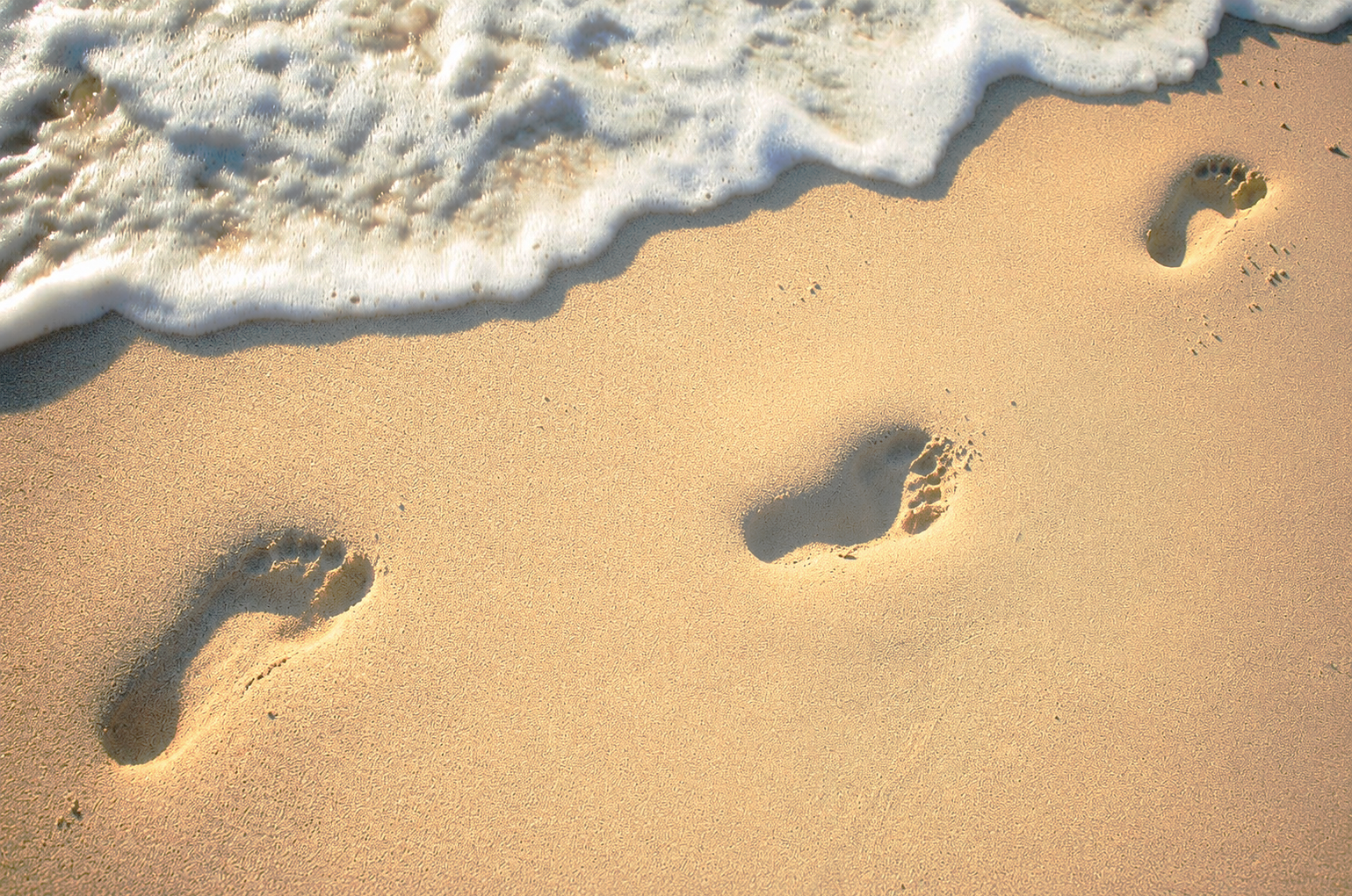 Footprints on the sandy shore near the water's edge with waves washing up on the beach.