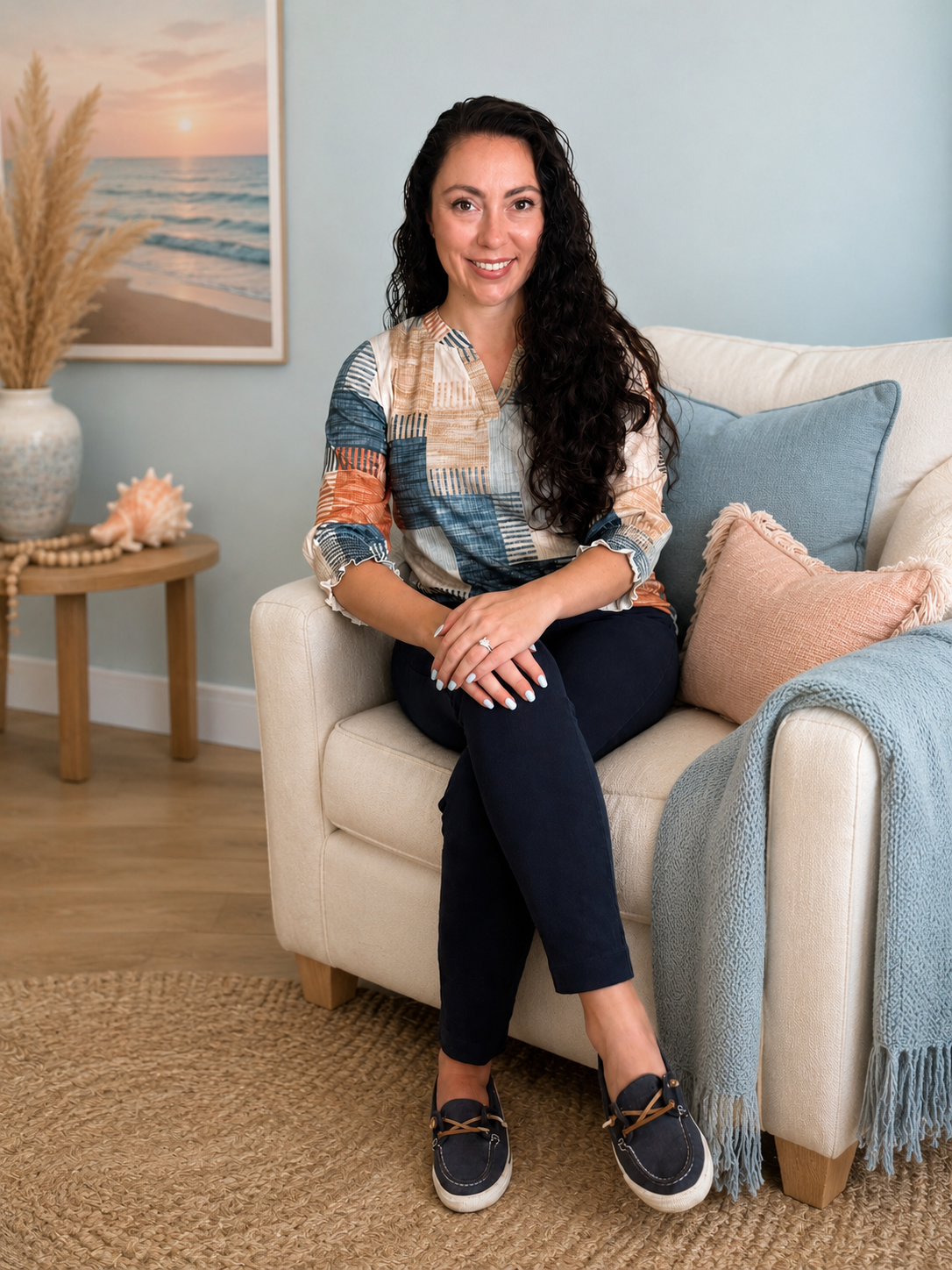 Smiling woman with long curly dark hair sitting on a beige sofa in a living room with beach-themed decor, including a framed picture of a sunset over the ocean, a vase with pampas grass, and seashells on a side table.