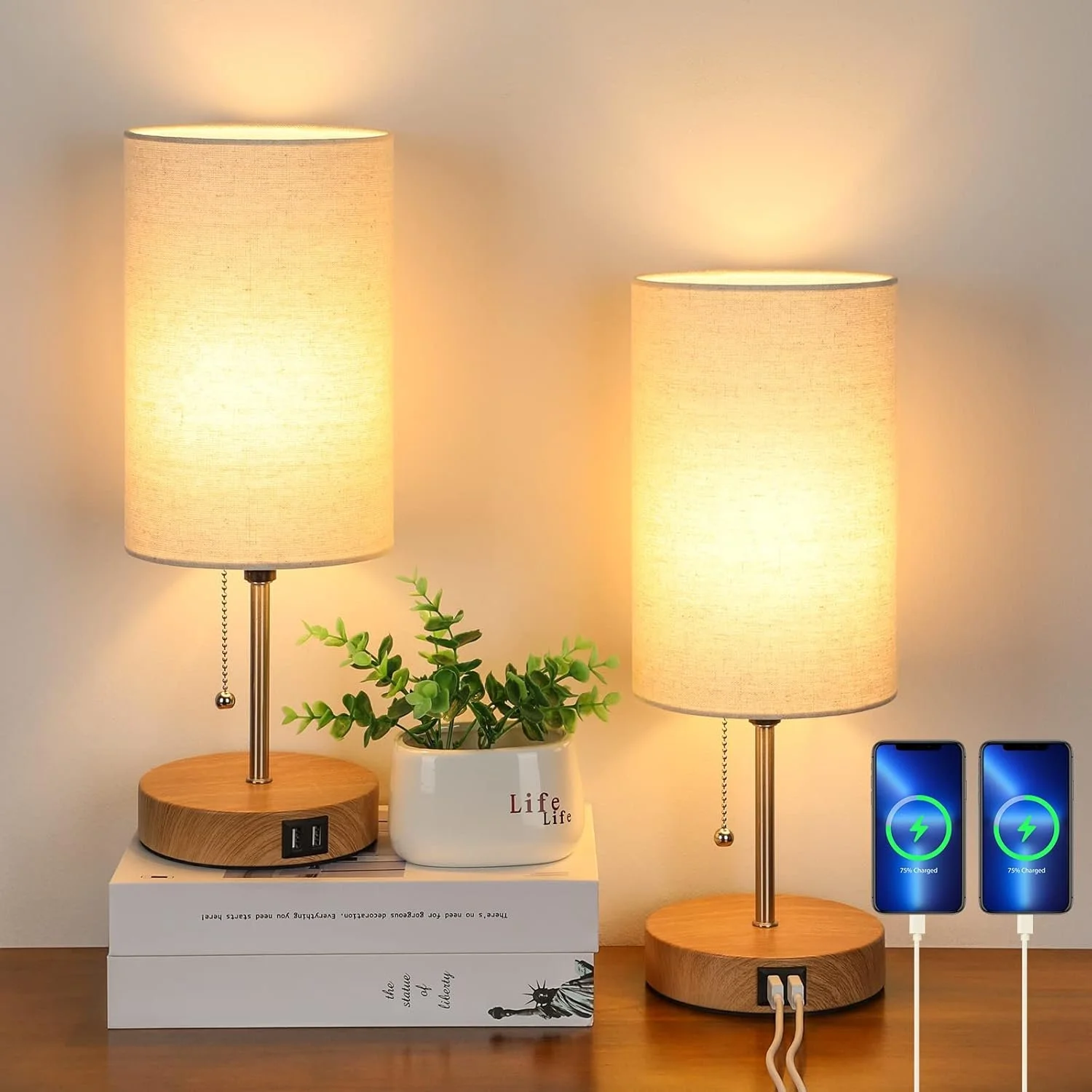 Two beige table lamps with wooden bases on a wooden surface, a white pot with green foliage, a large white book, and two smartphones charging with green battery icons, arranged on a side table against a plain wall.