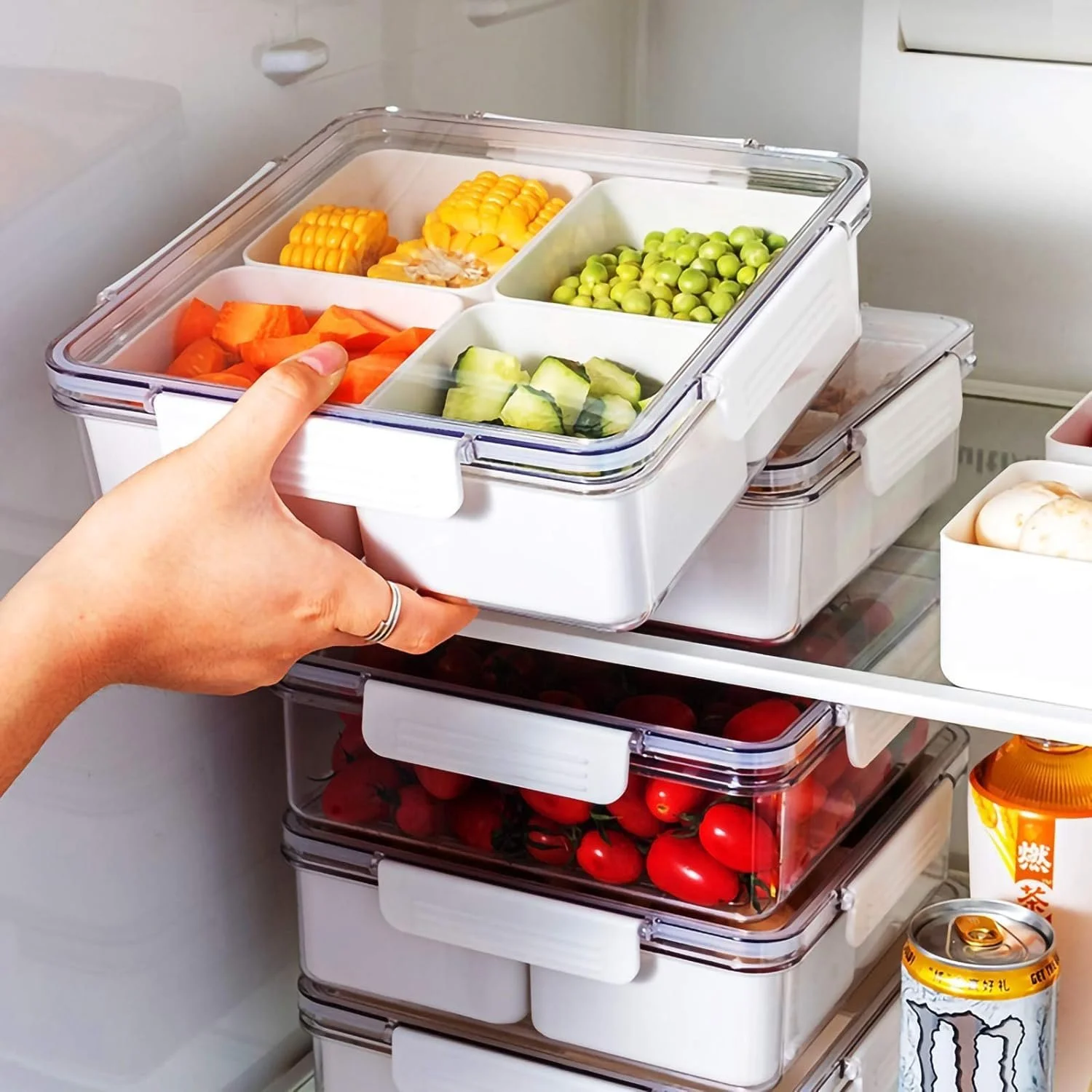 Close-up of a hand reaching into a clear plastic food storage container with sections filled with corn on the cob, green peas, chopped cucumbers, and sliced carrots. The container is on a shelf with additional stacked containers with tomatoes, eggs, and drinks beside it.