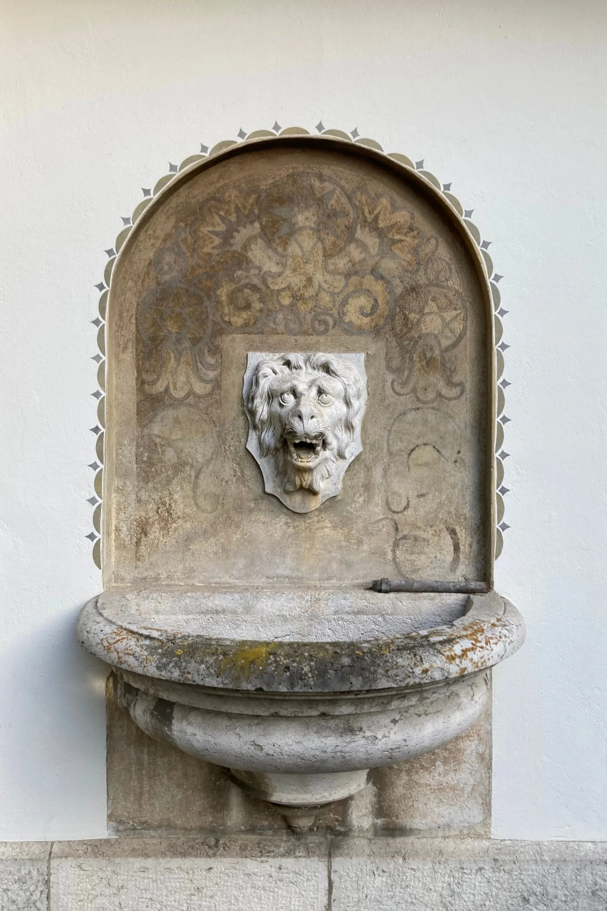 Steinerne Brunnen mit einer Löwen-Relief am Rand, befindet sich an einer weißen Wand.
