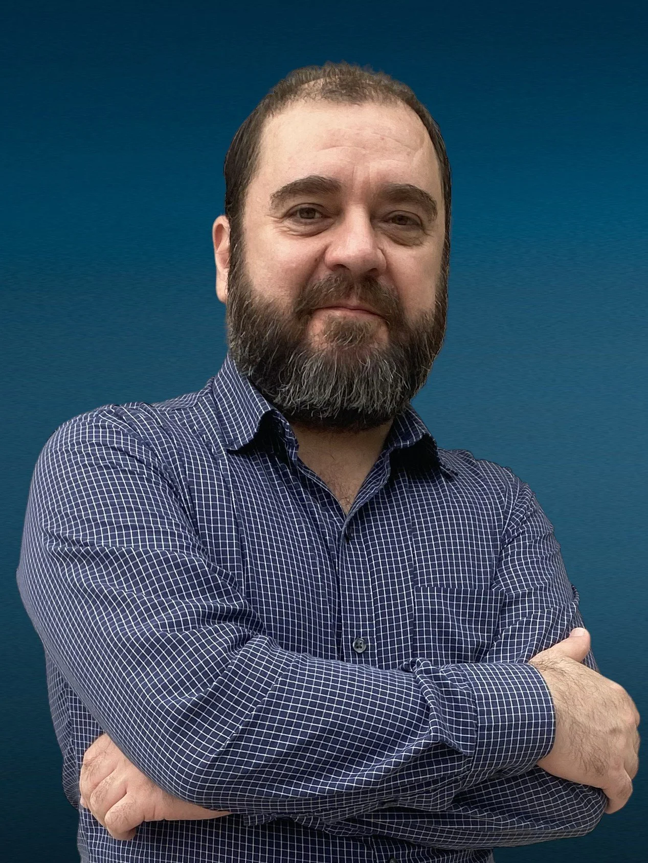 A man with a beard and mustache wearing a checkered blue shirt standing with arms crossed against a blue background.