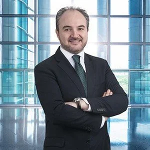 A man in a business suit standing with arms crossed in front of a large glass window with a modern building behind him.