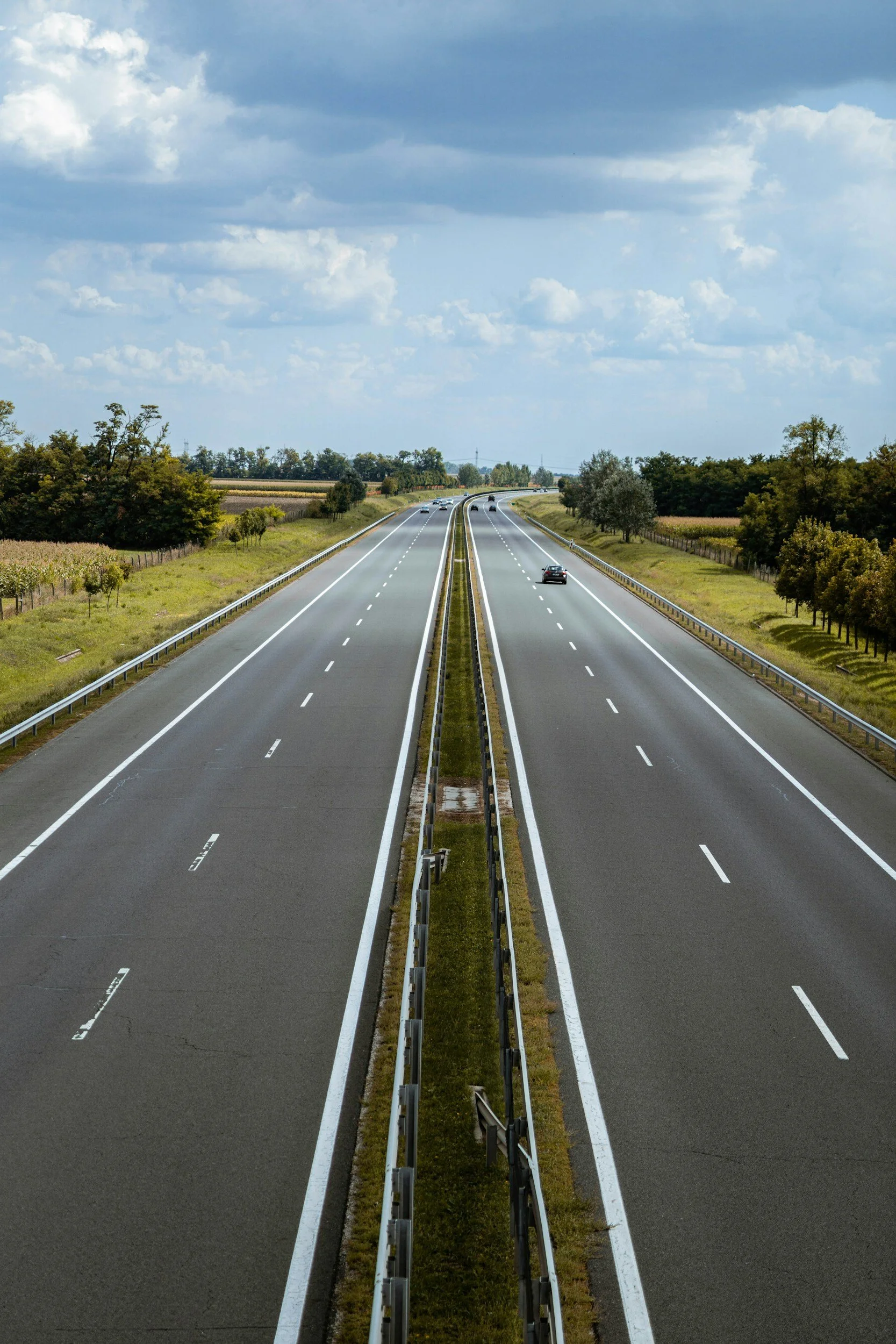 A highway with multiple lanes stretching into the distance, flanked by green fields and trees under a partly cloudy sky.