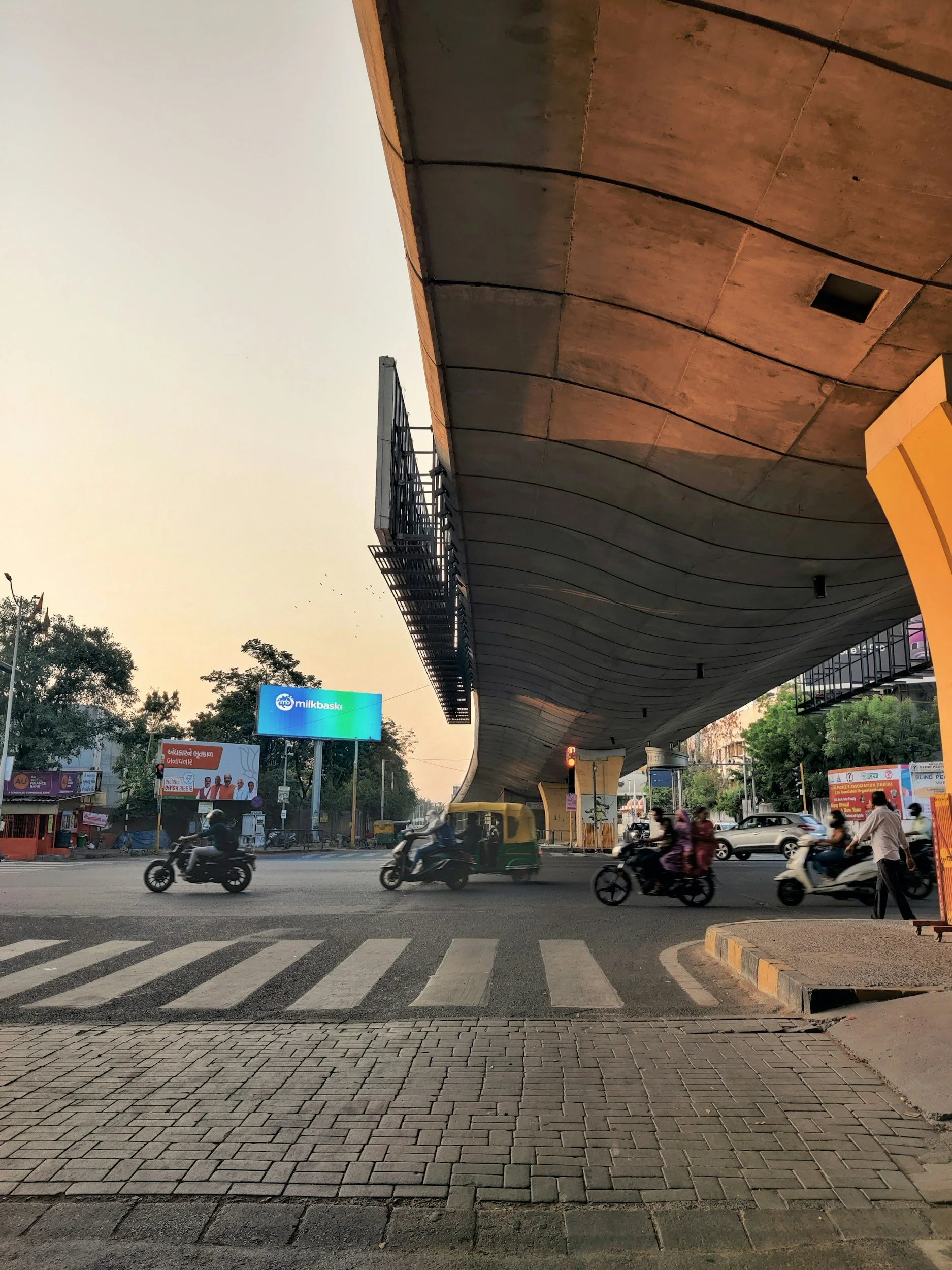 This image shows a busy street scene with motorcycles and a few pedestrians crossing at a zebra crossing. There's a large modern building with an overhanging roof structure and a billboard advertising 'Milk Baska' on the left side. The sky is clear, indicating early evening or late afternoon.