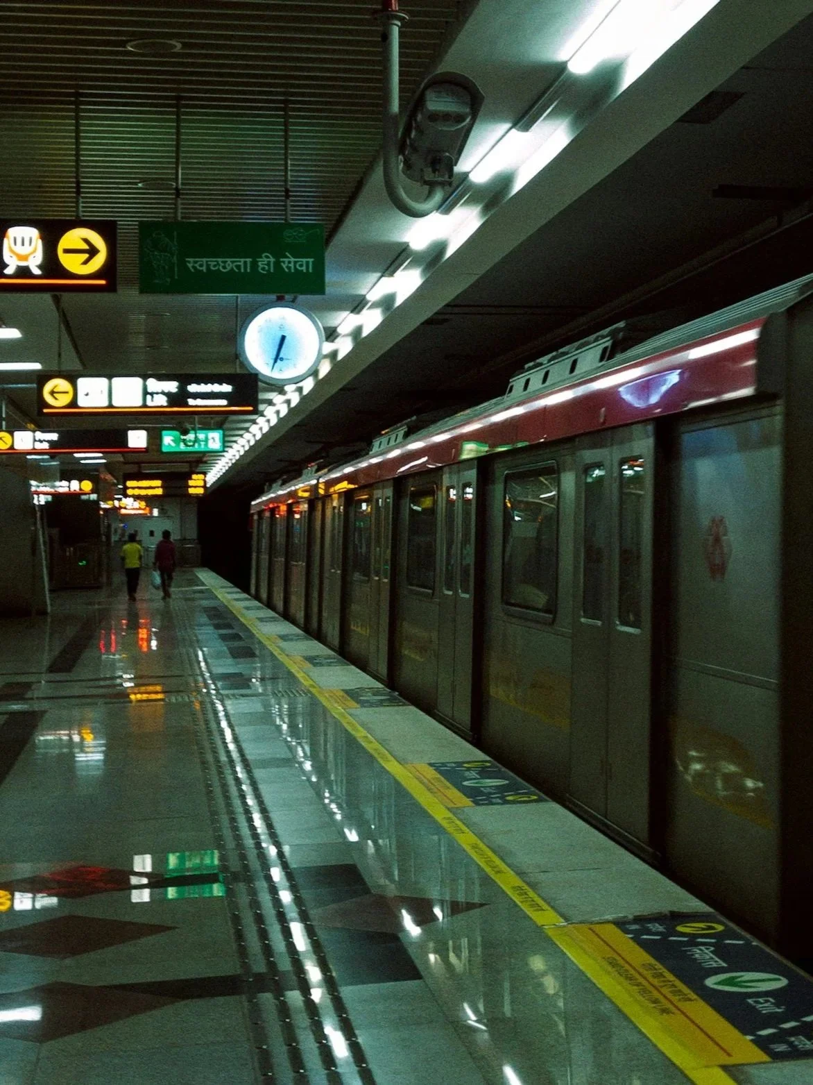 An underground metro train platform with a train on the right side, a clock, and illuminated signs with directions. Two people walk along the platform toward an exit.