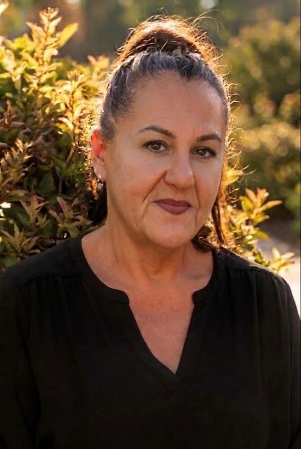 Close-up of a woman with dark hair in a high ponytail, wearing dark lipstick and a black top, standing outdoors with bushes and sunlight in the background. Coach