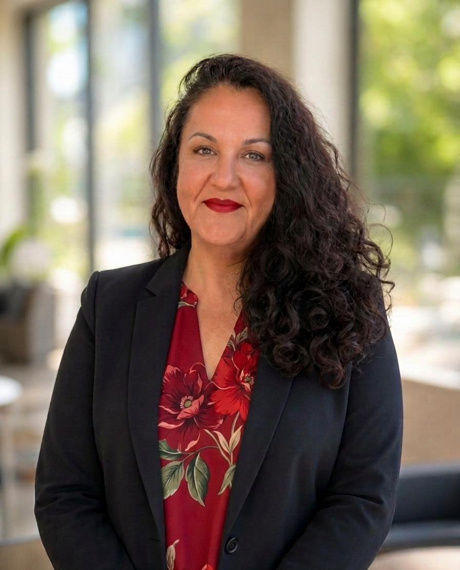 A woman with long curly dark hair and red lipstick, wearing a black blazer over a red floral blouse, standing indoors with large windows and greenery in the background.