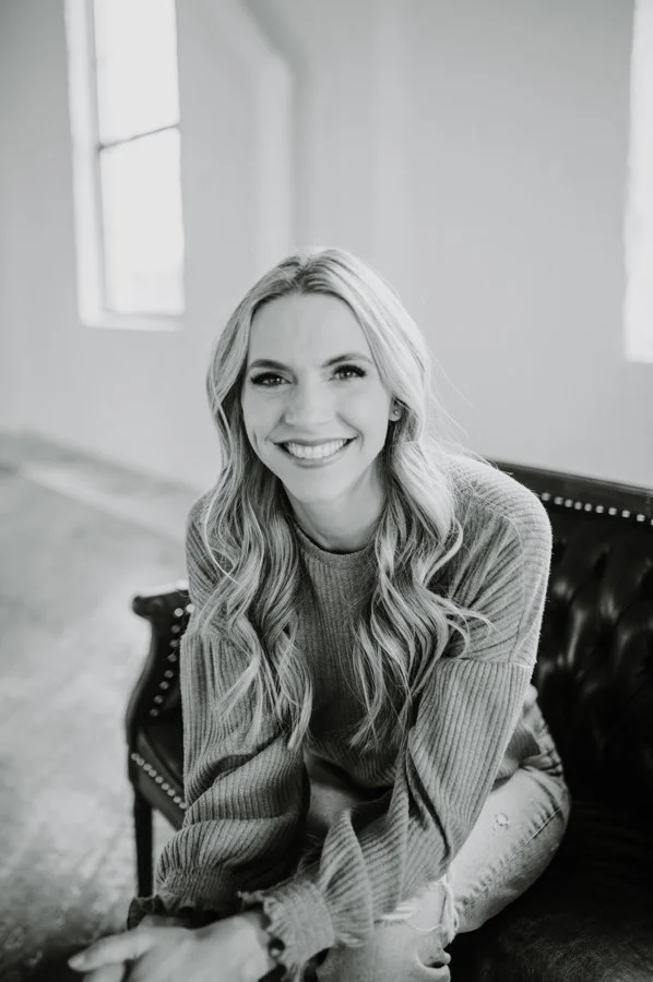 A woman with long wavy hair smiling, sitting on a vintage black leather couch in a bright room with large windows.