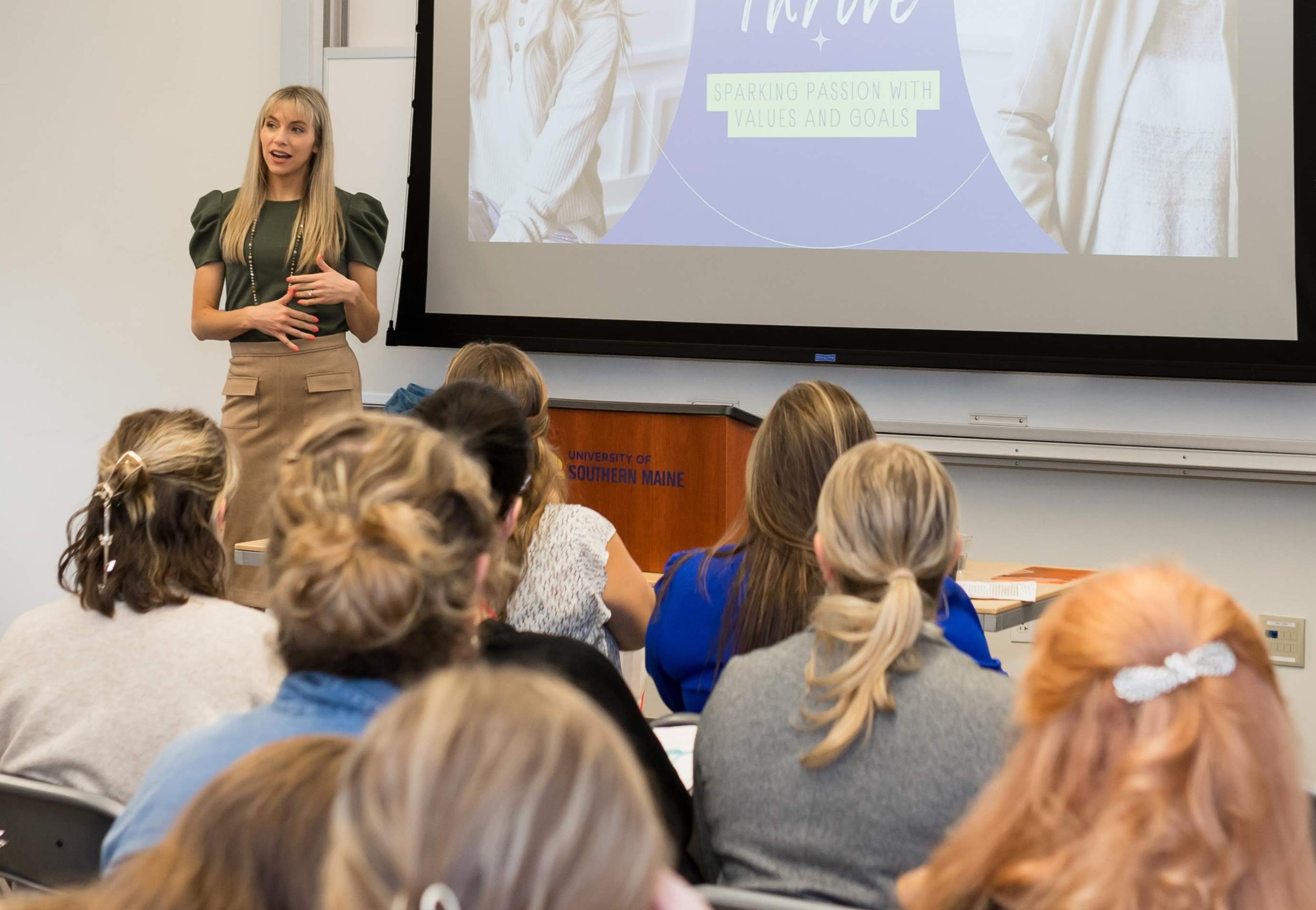 A woman giving a presentation in a classroom with students seated and facing her. The presentation slide on the screen reads 'SPARKING PASSION WITH VALUES AND GOALS'. The podium has an inscription of 'UNIVERSITY OF SOUTHERN MAINE'.